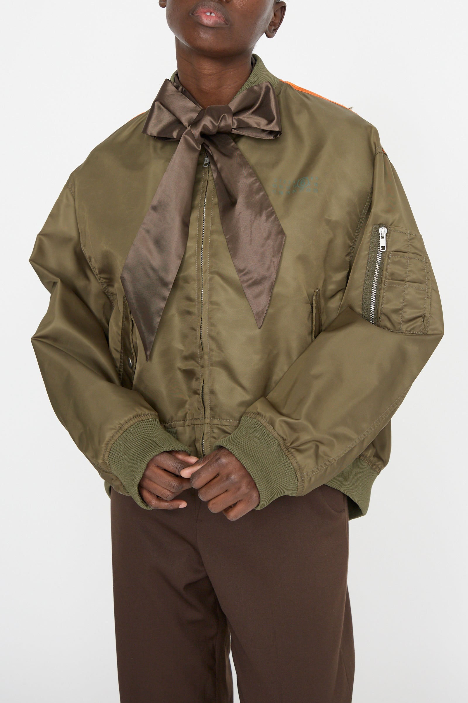 A person is wearing a lightweight MM6 Sports Jacket in Military, featuring a large brown bow at the neck, paired with brown pants against a plain background.