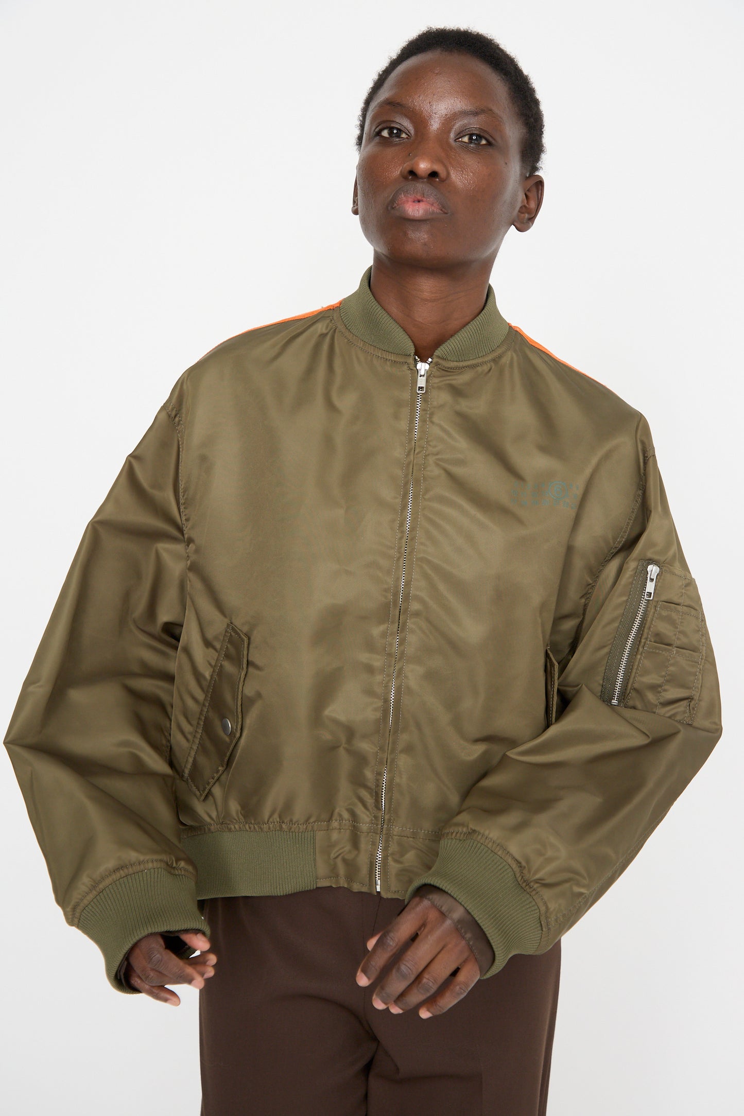 A person wears an olive green MM6 Sports Jacket, crafted in lightweight polyamide with orange accents and a color-block panel design, paired with brown pants, standing against a plain white background.