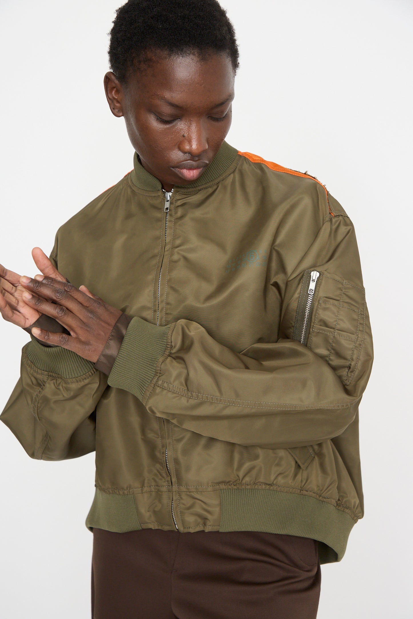 A person models the MM6 Sports Jacket, an olive green, lightweight bomber with a color-block panel design and vibrant orange accents. It's paired with brown pants as they stand looking down with their hands clasped.