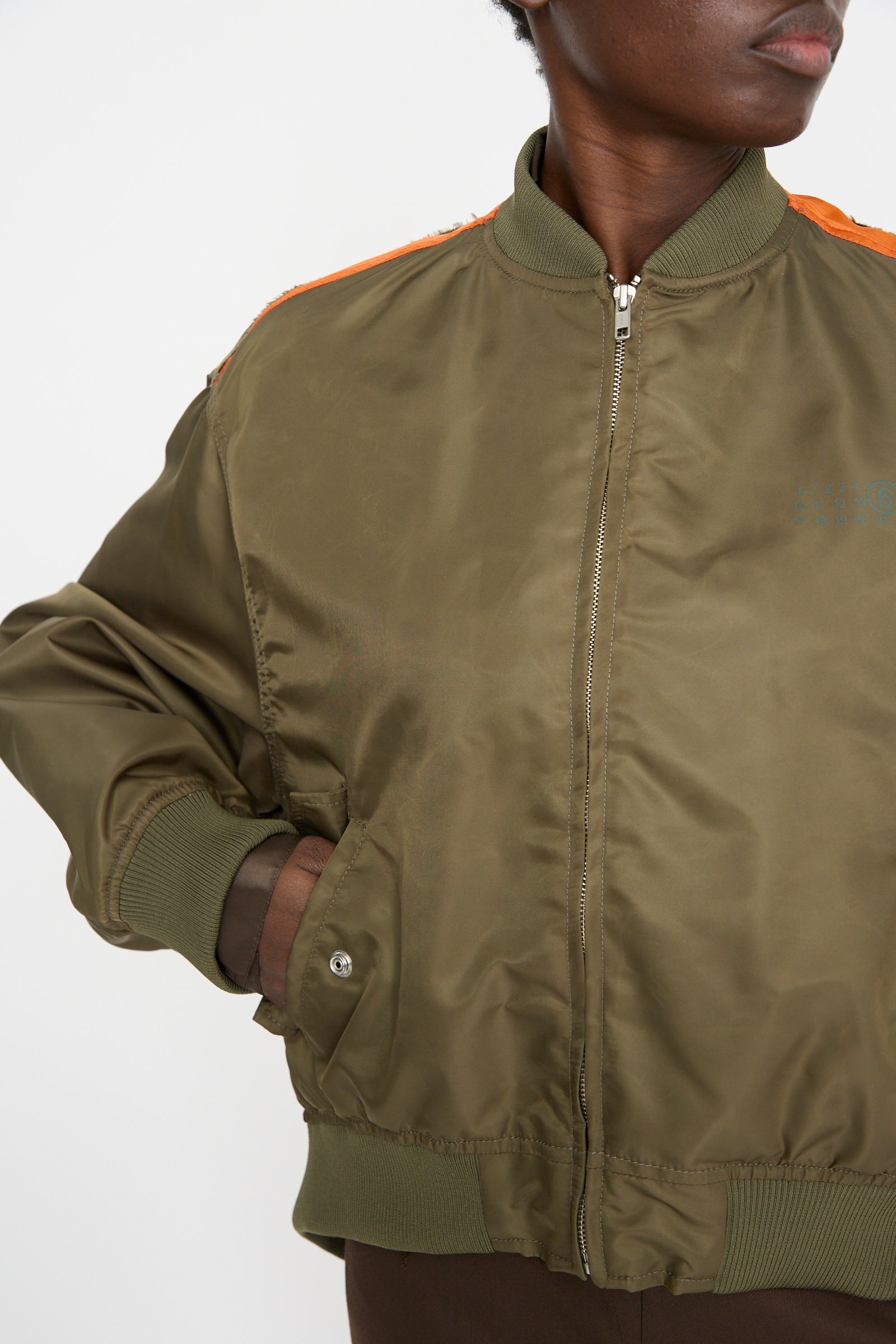 A person is wearing an MM6 Sports Jacket, a stylish olive green bomber with orange accents and hands in pockets. Made from durable olive polyamide, this lightweight jacket boasts a trendy color-block design that modernizes the classic look.