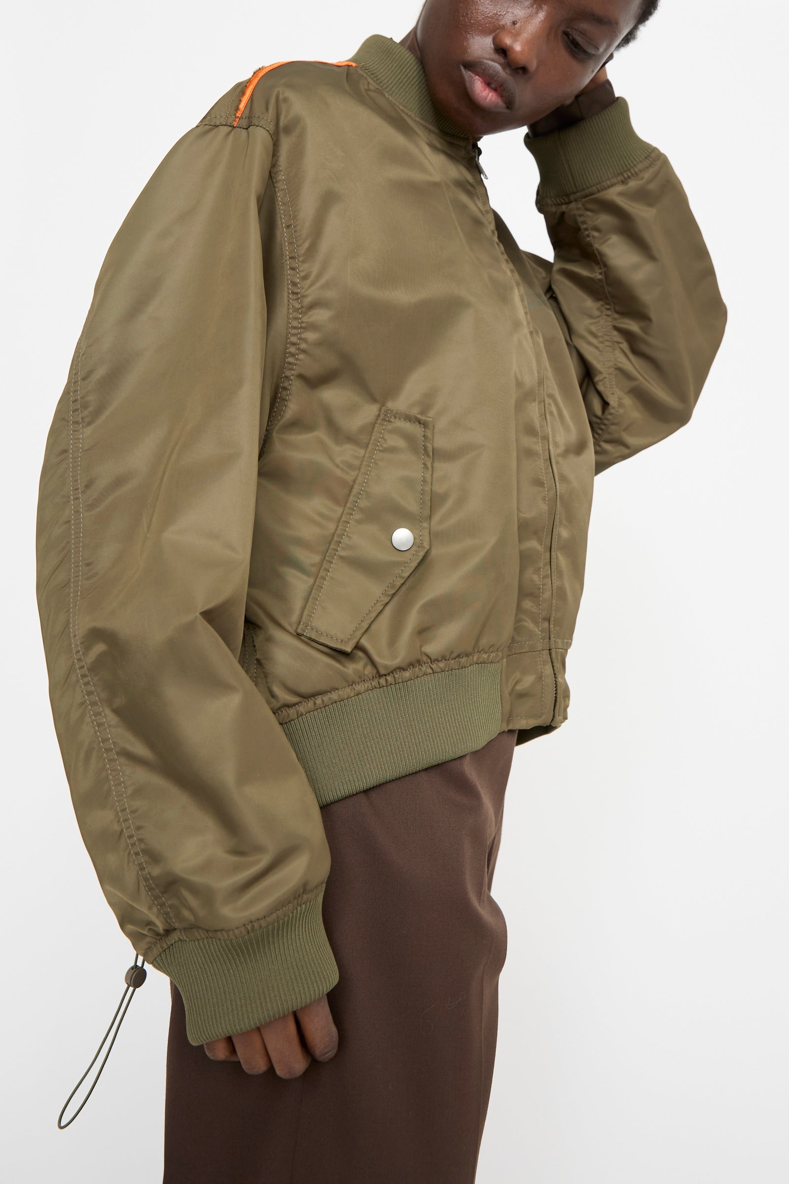 Someone is wearing an olive green MM6 Sports Jacket in Military with orange lining and dark brown pants. The lightweight polyamide jacket, featuring a unique color-block panel, has one hand raised to adjust the collar.