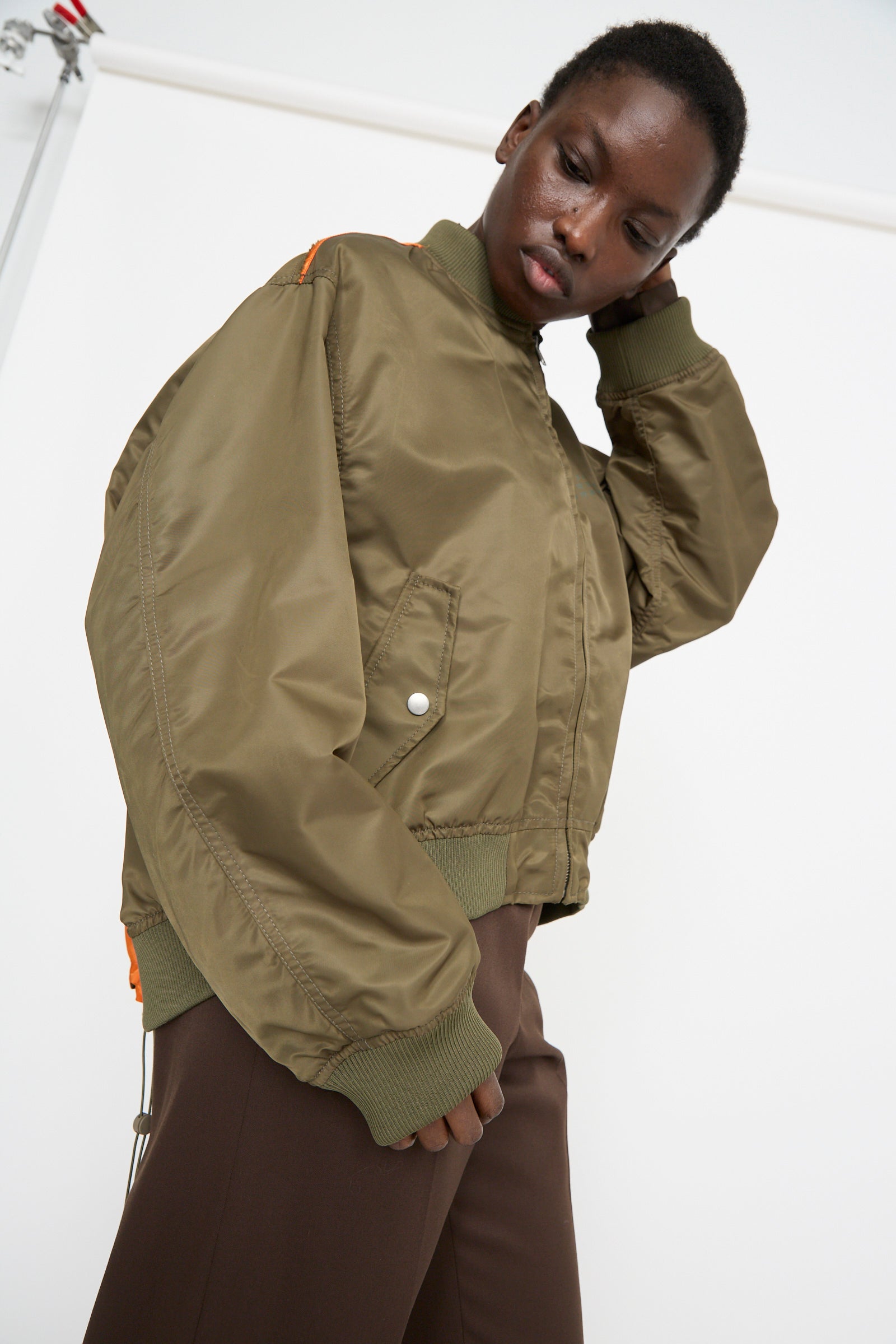 A person wearing an MM6 Sports Jacket in Military with color-block panels and brown pants stands with one hand on their head against a light background.