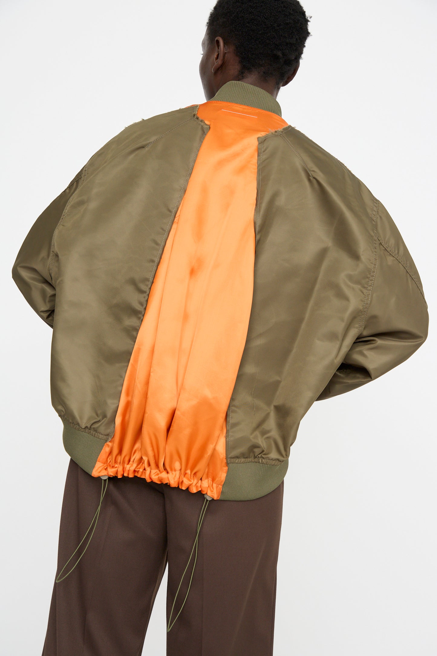 A person is wearing an oversized MM6 sports jacket made of olive polyamide with an orange lining and brown pants, featuring a subtle color-block panel design, while facing away.