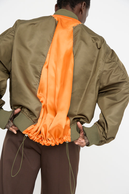 A person stands against a white background wearing an olive polyamide MM6 Sports Jacket with orange lining and stylish color-block panels, paired with brown pants.