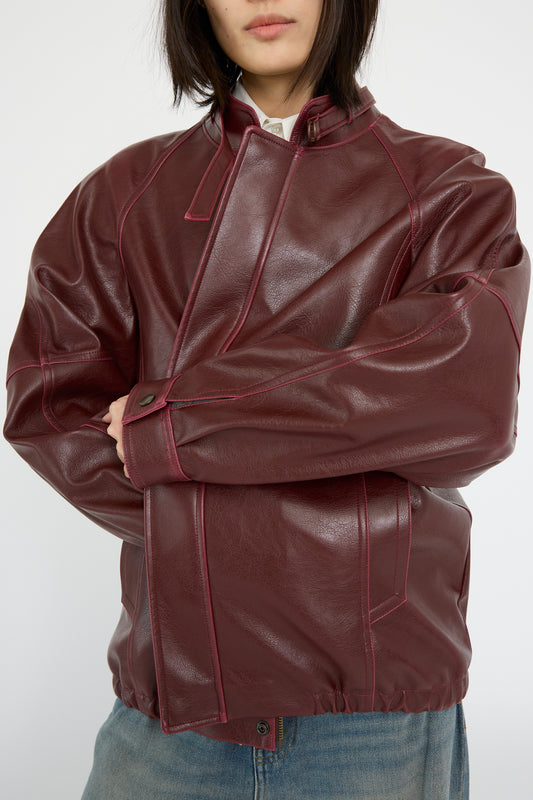 Against a plain background, someone wears an MM6 Sports Jacket in burgundy faux-leather, featuring a relaxed fit with a high collar and buttoned cuffs.