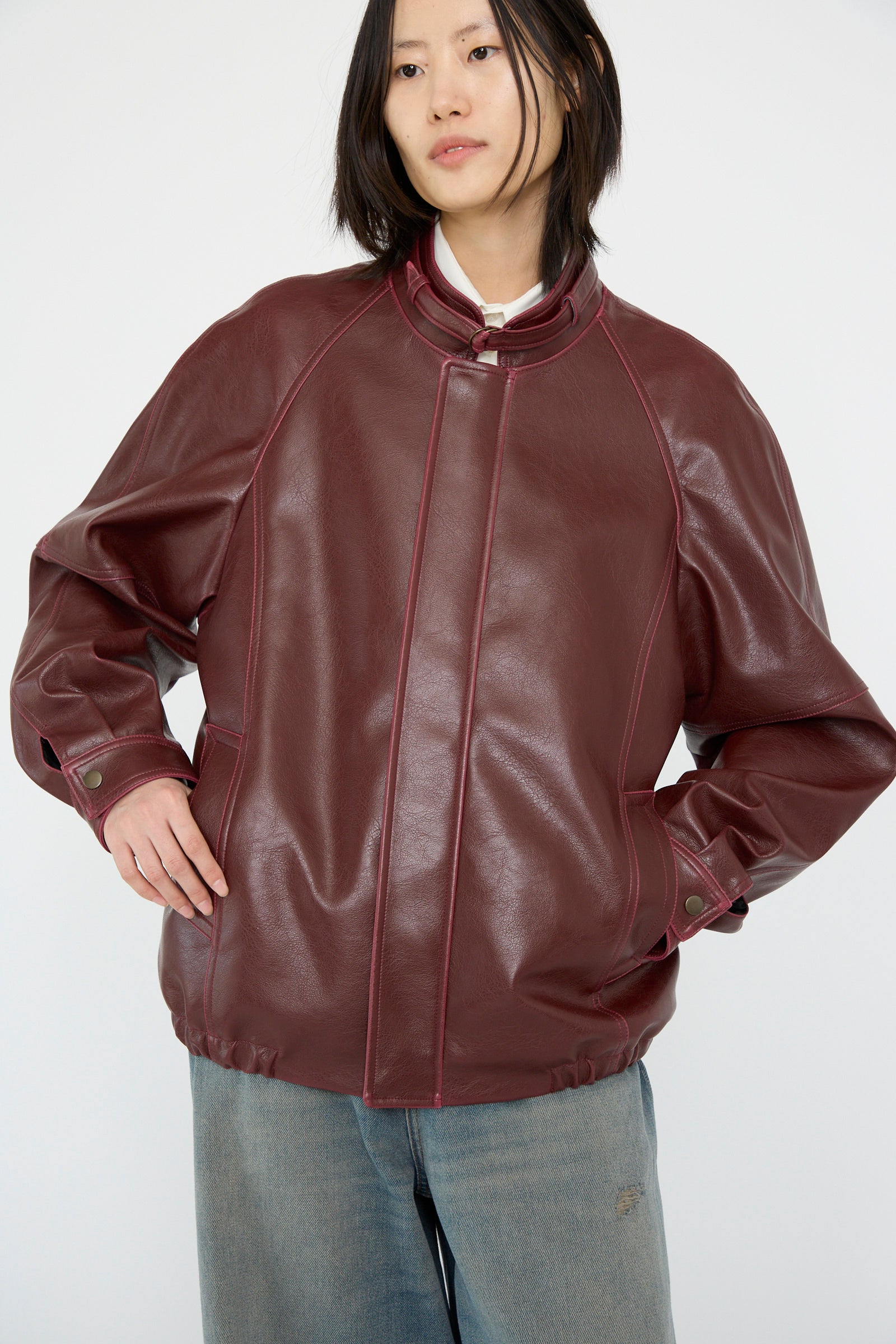 A person wearing an MM6 Sports Jacket in burgundy faux-leather with a snap button collar, paired with blue jeans, arms slightly bent.