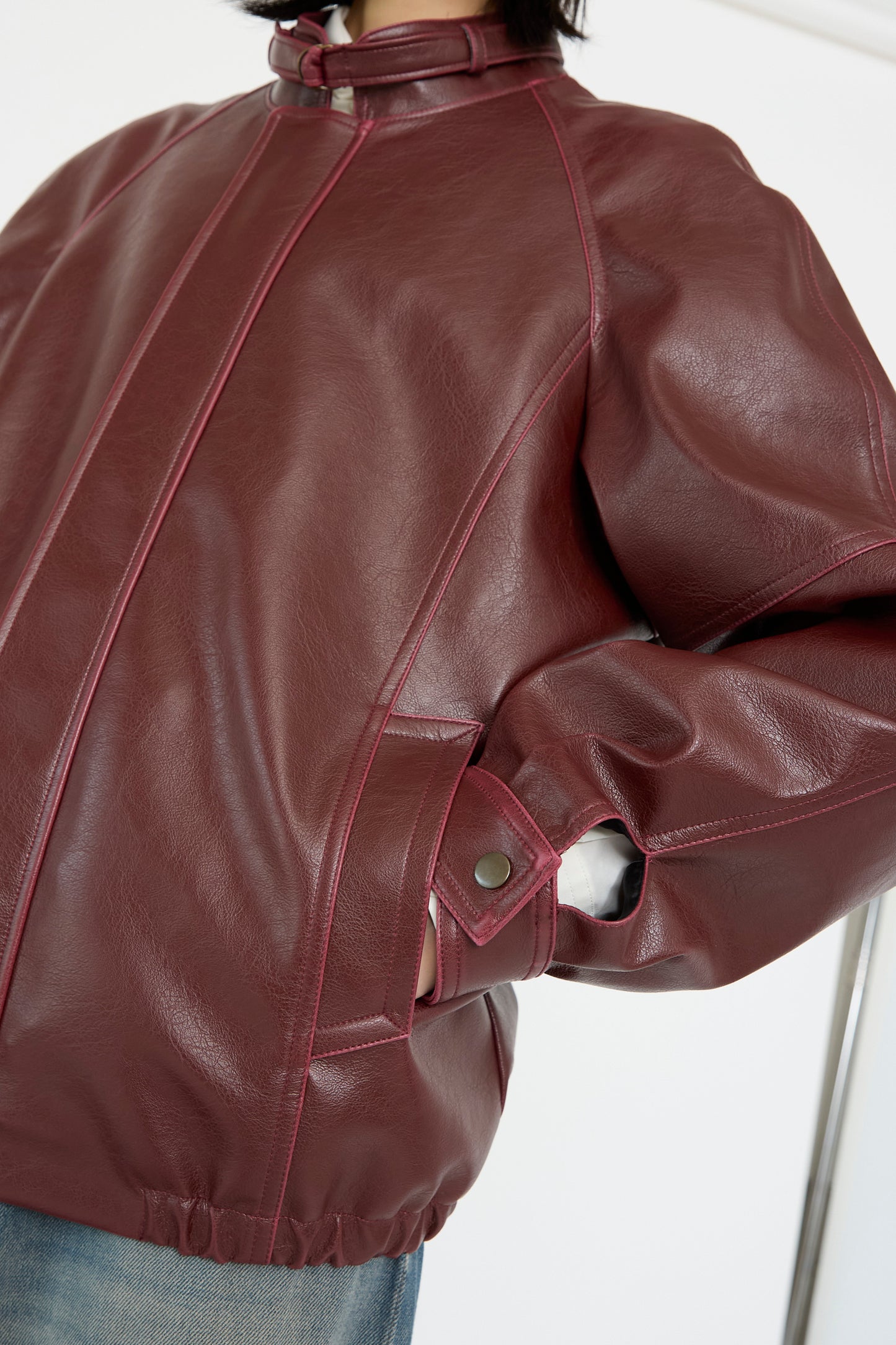 A person stands with hands in pockets, wearing a burgundy MM6 Sports Jacket made of faux leather, featuring pink stitching and metal snap buttons.