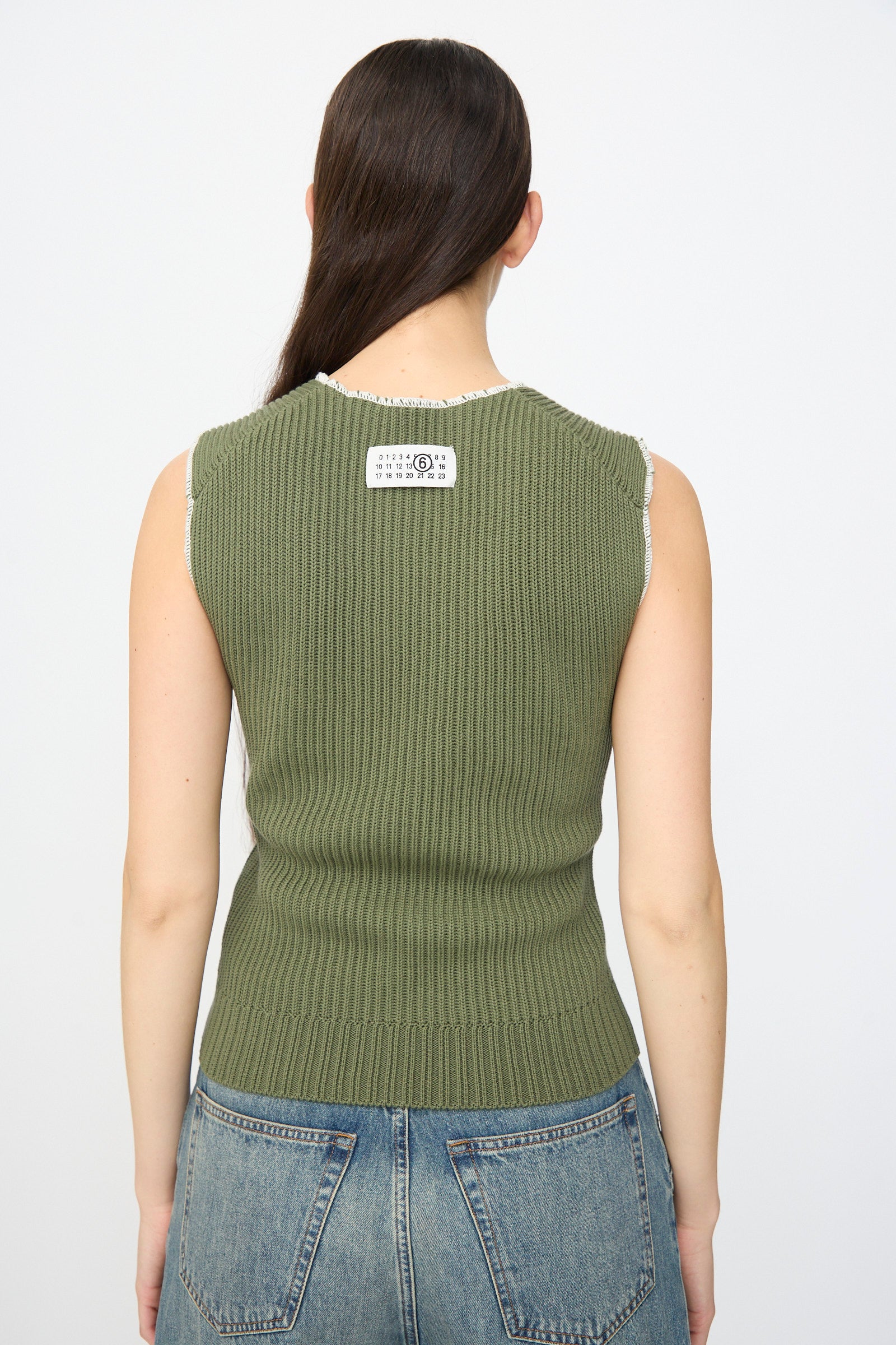 A person with long dark hair faces away from the camera, wearing the MM6 Top in Khaki, a slim fit ribbed knit cotton tank with a white label on the upper back, styled with blue jeans.