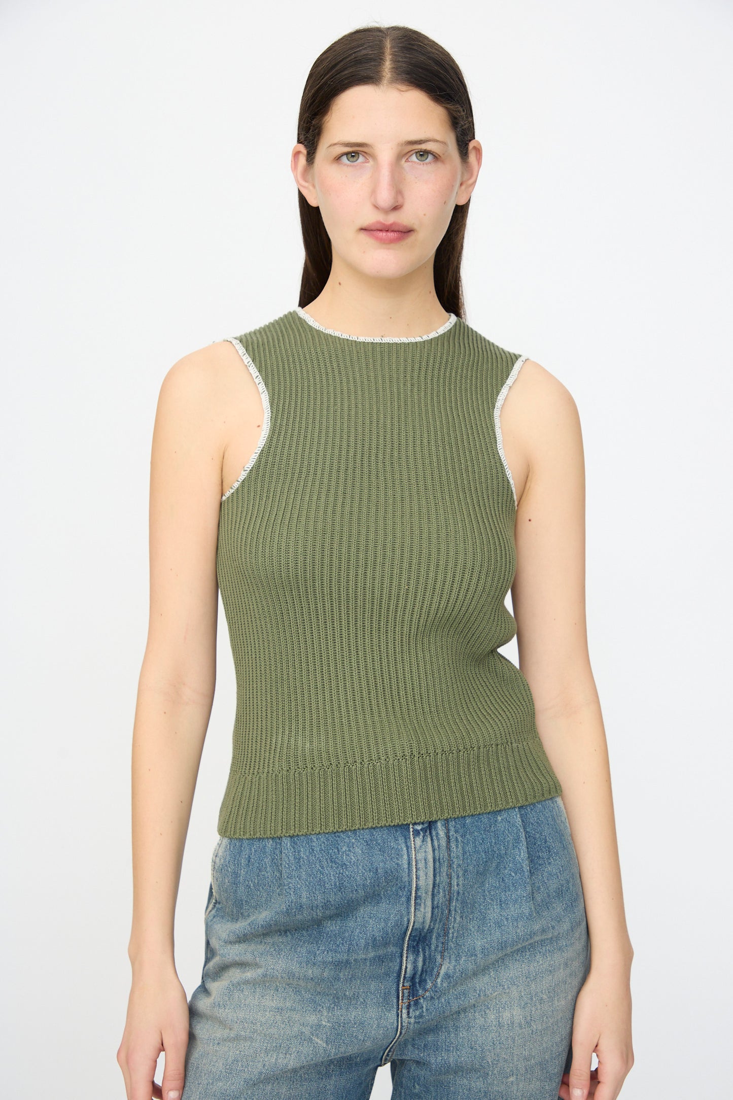 A woman with straight brown hair wears the MM6 Top in Khaki, a slim fit ribbed knit cotton tank, paired with blue jeans and standing against a plain white background.
