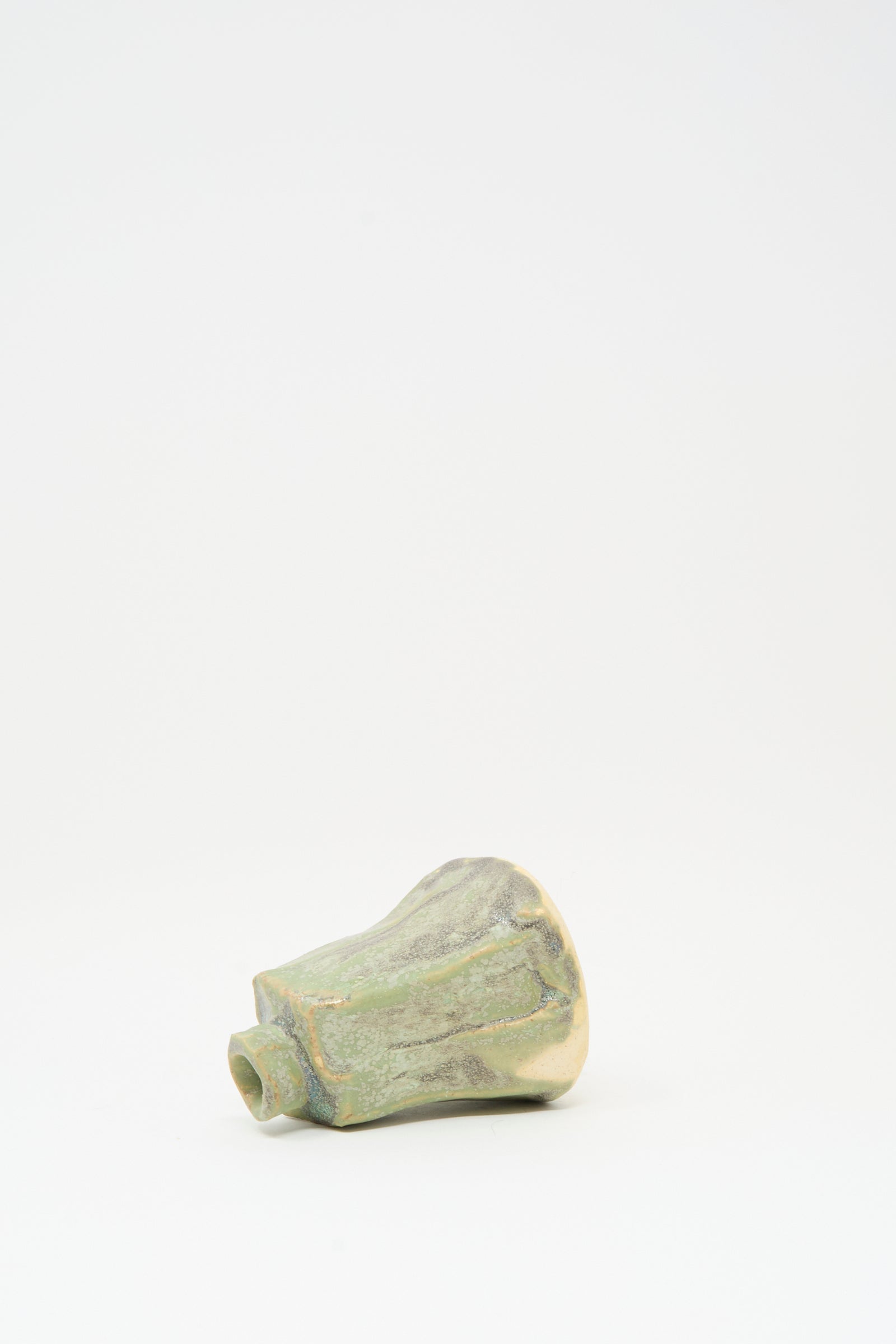 The MONDAYS Carved Bud Vase in Lichen Green, a small handmade ceramic piece with an asymmetrical, marbled surface, rests on its side against a plain white background.
