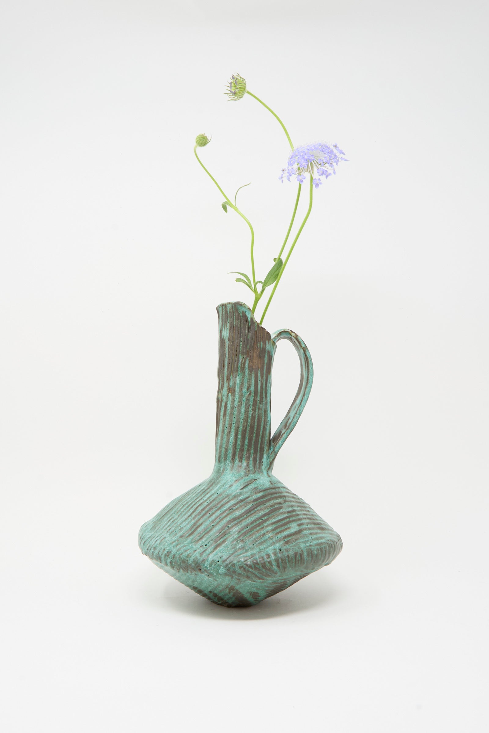 The MONDAYS Corrugated Pitcher in Green I, featuring a sculptural handle and textured glaze, holds two delicate purple flowers against a plain white background.