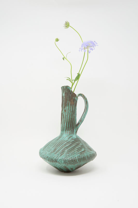 The MONDAYS Corrugated Pitcher in Green I, featuring a sculptural handle and textured glaze, holds two delicate purple flowers against a plain white background.