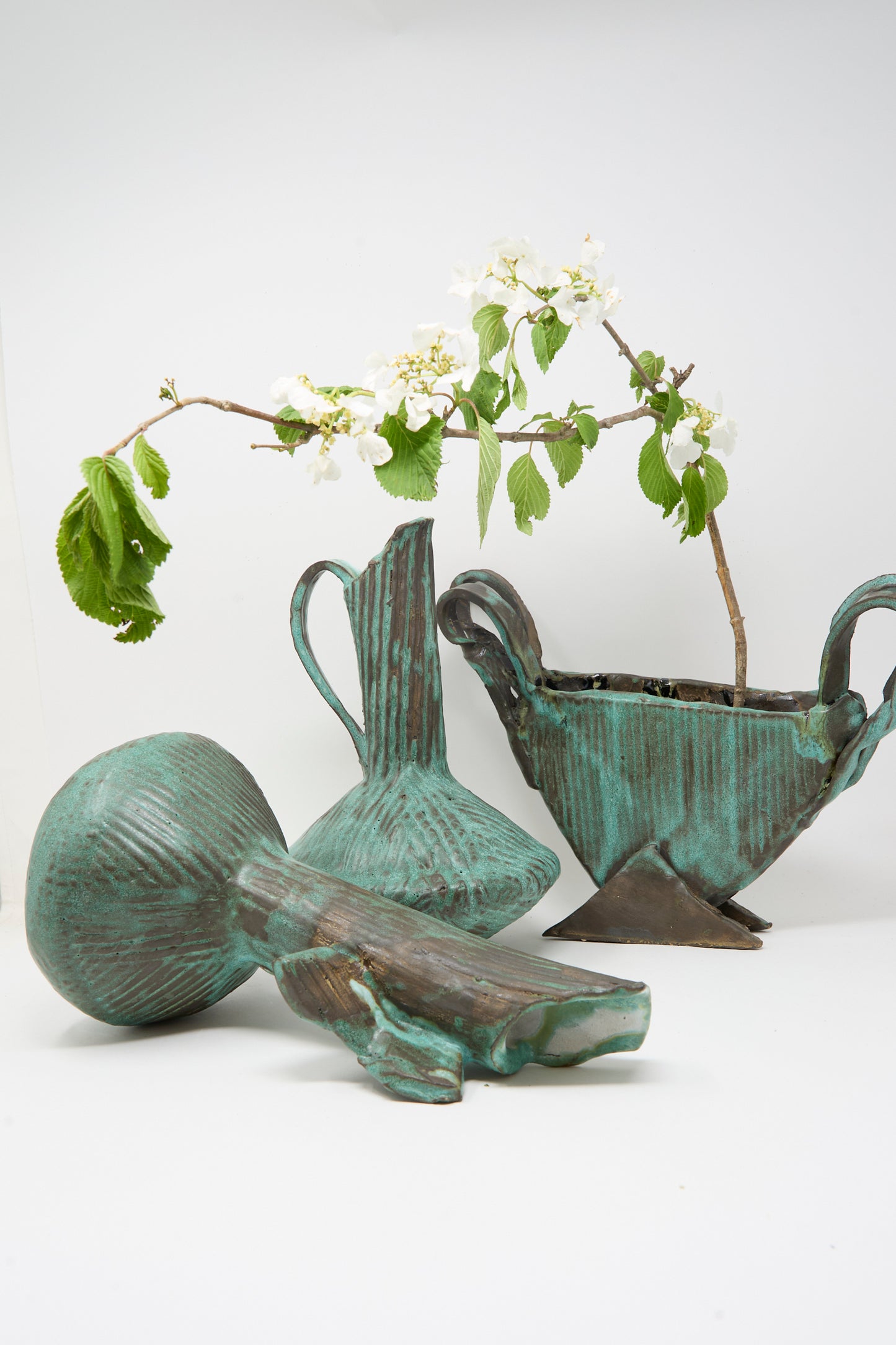 Three MONDAYS Corrugated Pitcher in Green I ceramic vessels are displayed on a white background; two hold branches with white flowers, while one sculptural pitcher rests on its side in the foreground.