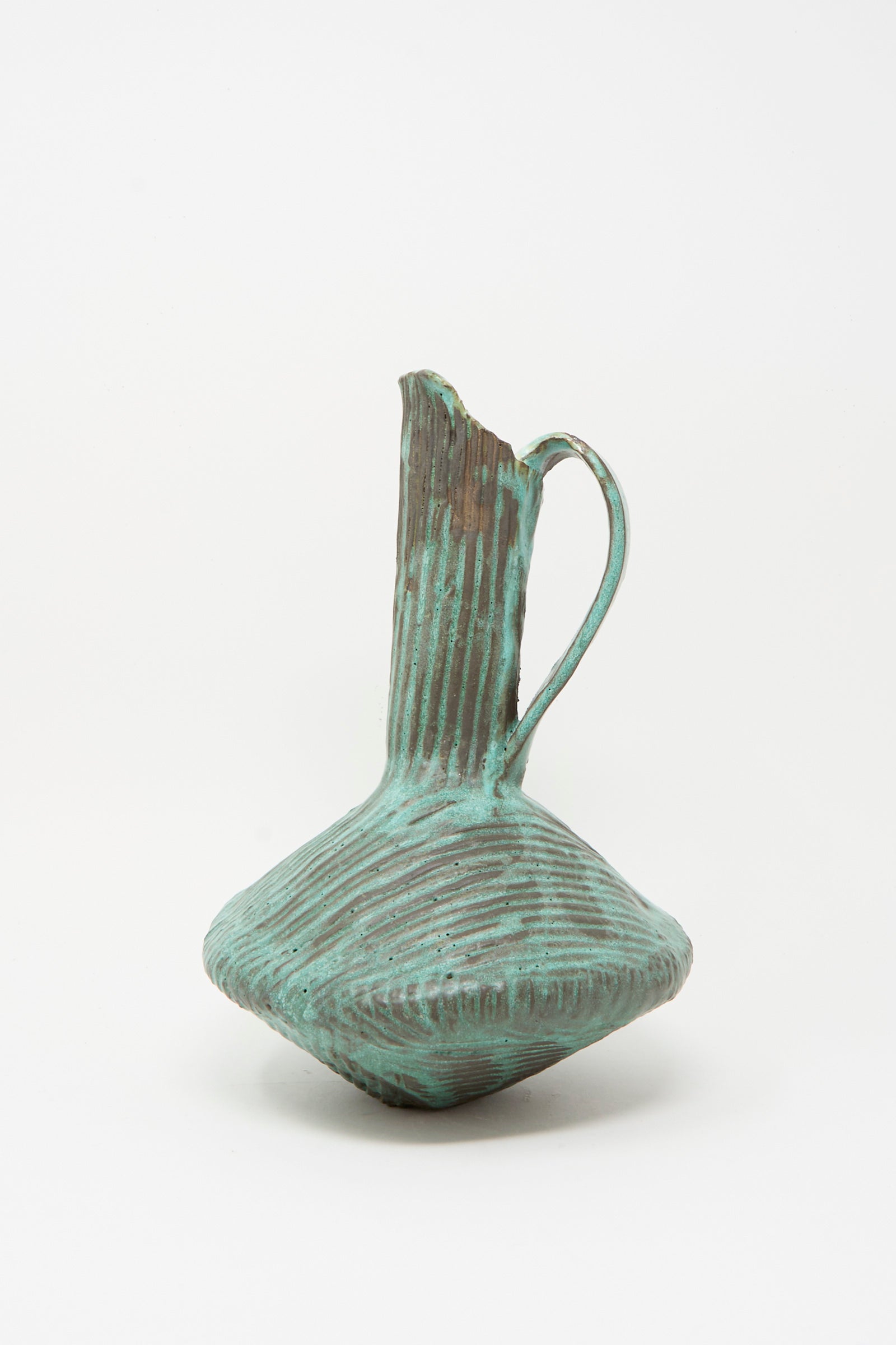 The Corrugated Pitcher in Green I by MONDAYS is a handmade ceramic piece featuring a ribbed texture, narrow neck, angular body, and single handle—a sculptural design in green against a plain white backdrop.