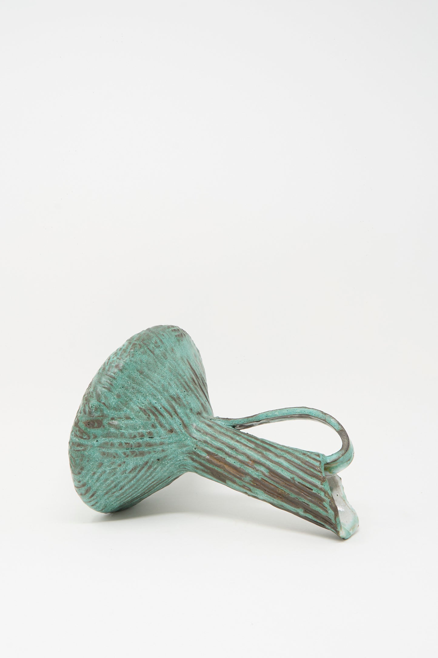 The Corrugated Pitcher in Green I by MONDAYS, a fluted sculptural piece with a handle and an ancient feel, rests on its side against a plain white background.