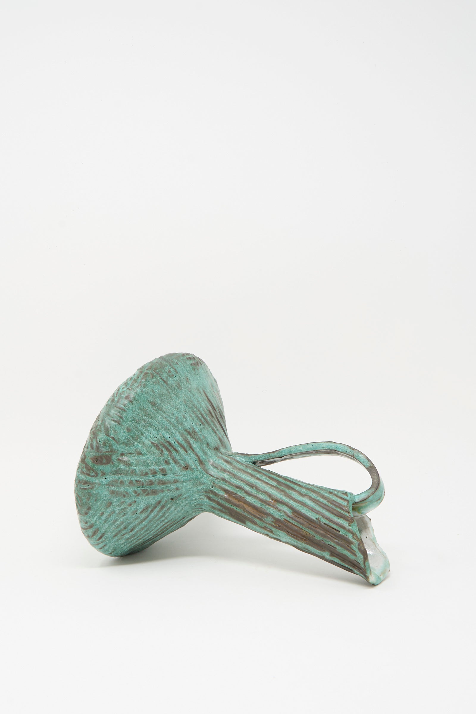 The Corrugated Pitcher in Green I by MONDAYS, a fluted sculptural piece with a handle and an ancient feel, rests on its side against a plain white background.