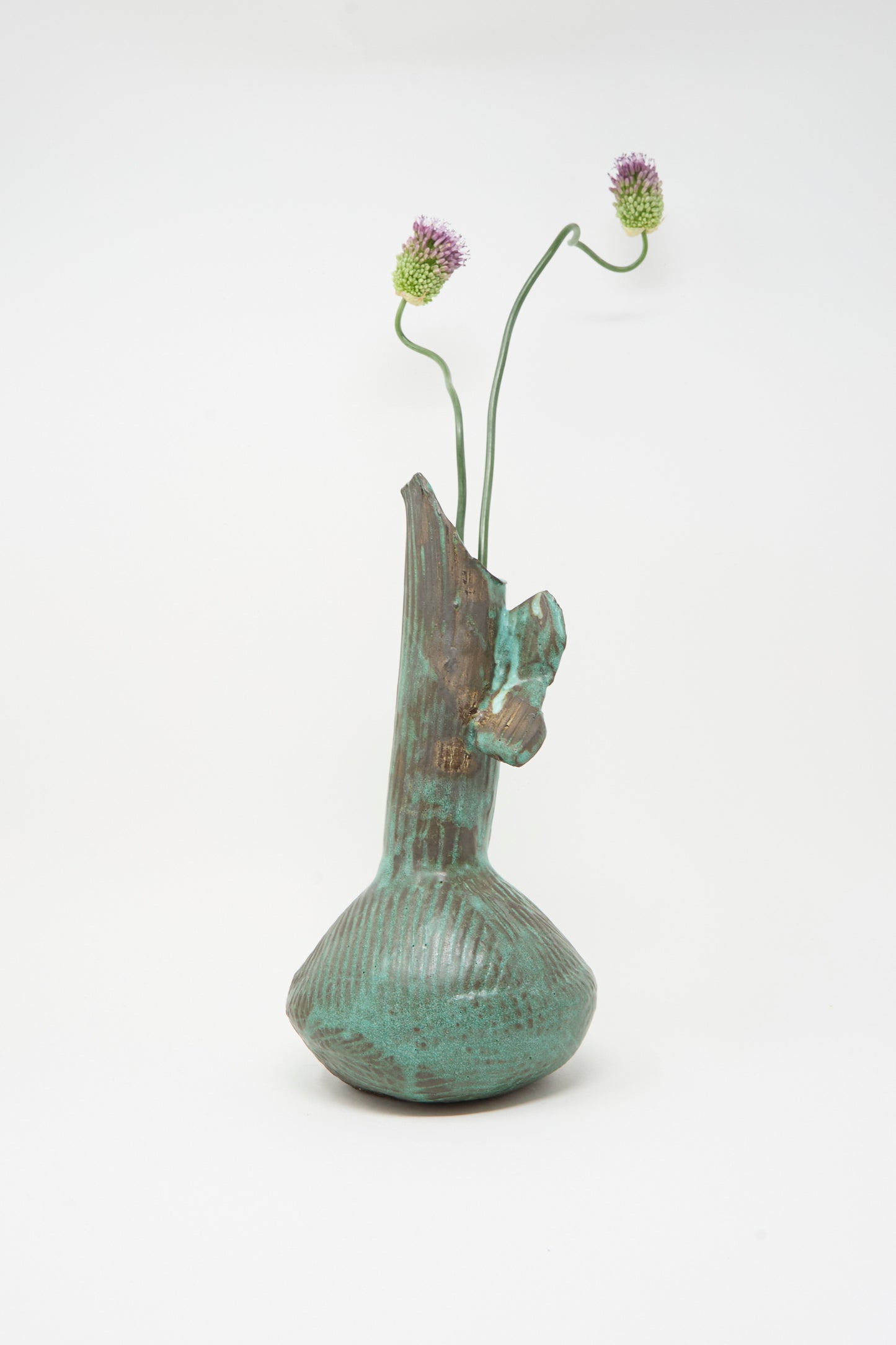 The MONDAYS Corrugated Pitcher in Green II, with its textured surface and asymmetrical Brooklyn-inspired design, holds two long-stemmed purple thistle flowers against a plain white background.