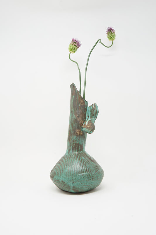 The MONDAYS Corrugated Pitcher in Green II, with its textured surface and asymmetrical Brooklyn-inspired design, holds two long-stemmed purple thistle flowers against a plain white background.
