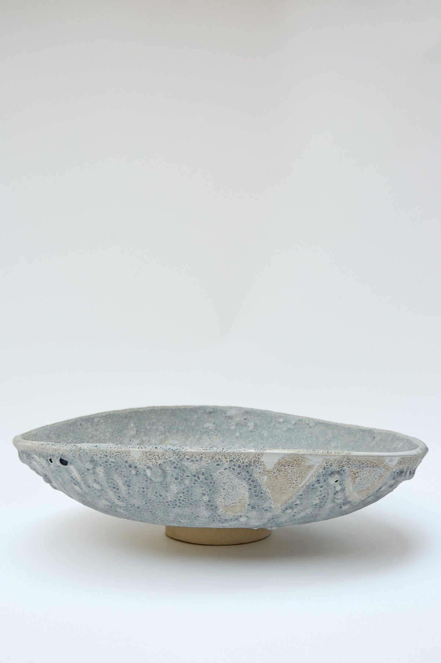 A round, shallow stoneware bowl with a gray marbled glaze, displayed against a plain white background. The textured lava glaze adds an intriguing touch to its minimalist design, the Lava Pedestal Bowl in Glazed Stoneware by MONDAYS.