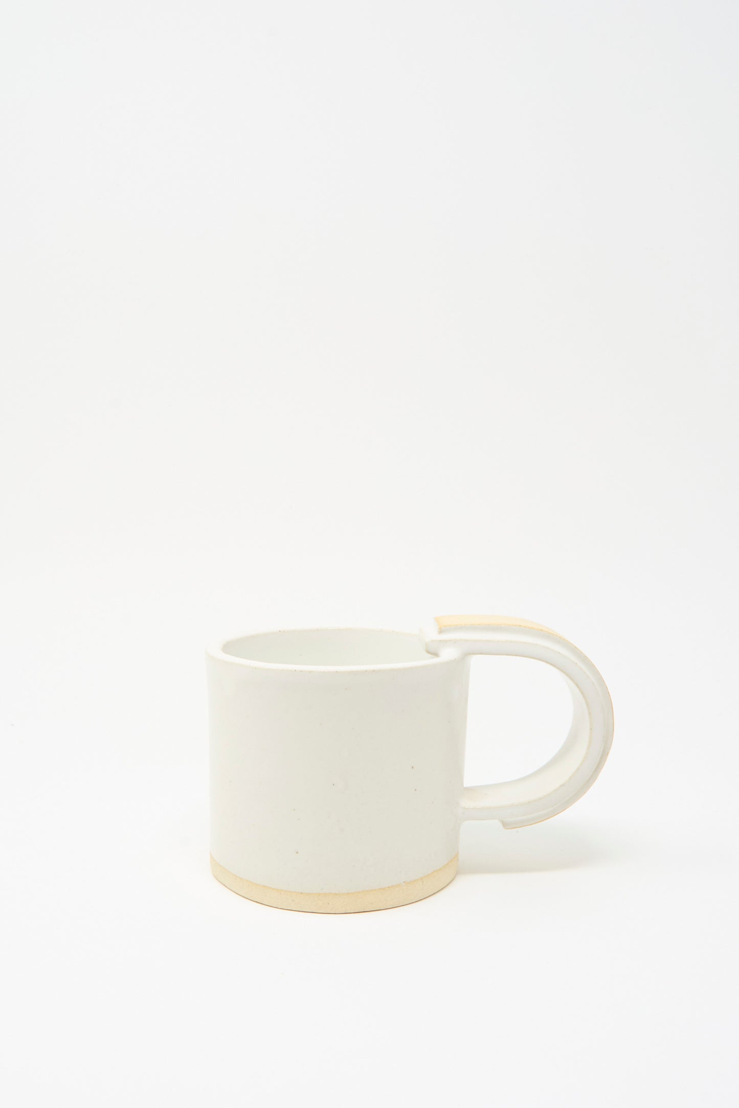 The MONDAYS Layered Mug in White on Sand features a handmade, slab-built design with a white ceramic body, rounded handle, and unglazed beige base, shown against a white background.