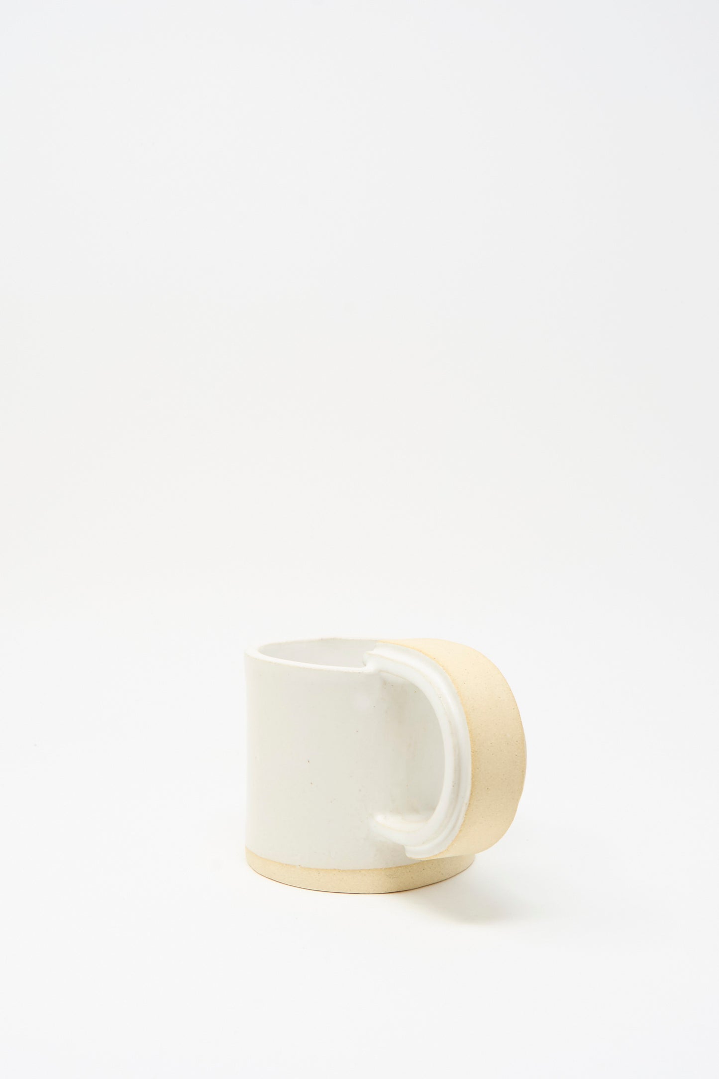 The MONDAYS Layered Mug in White on Sand features a handmade, slab-built design with a beige unglazed handle and base, and a white ceramic body. It’s photographed on a plain white background.