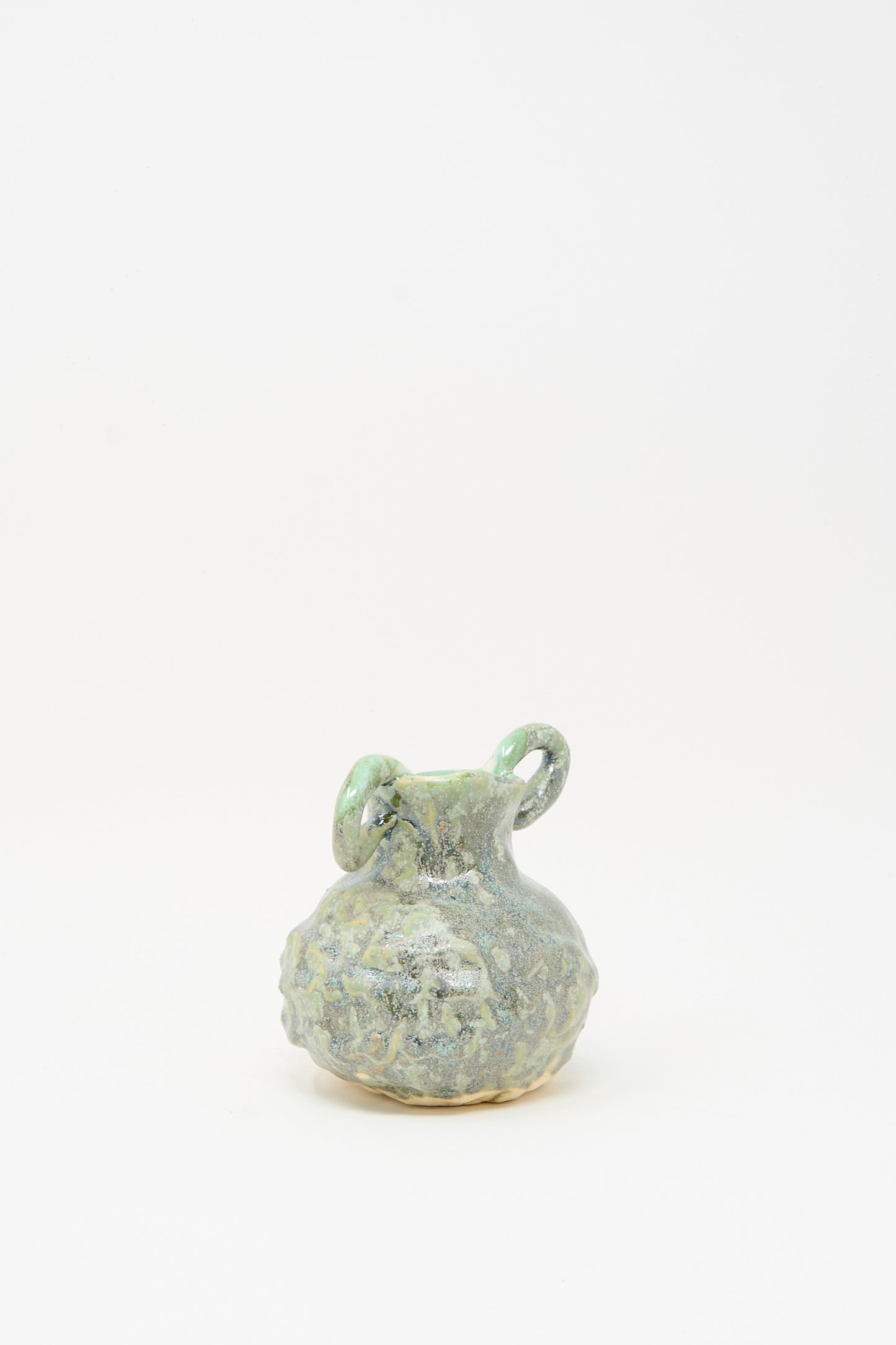 The MONDAYS Pinched Vase with Handles in Green features a small, round handcrafted design with two looped handles and a mottled green and gray glaze, shown against a plain white background.