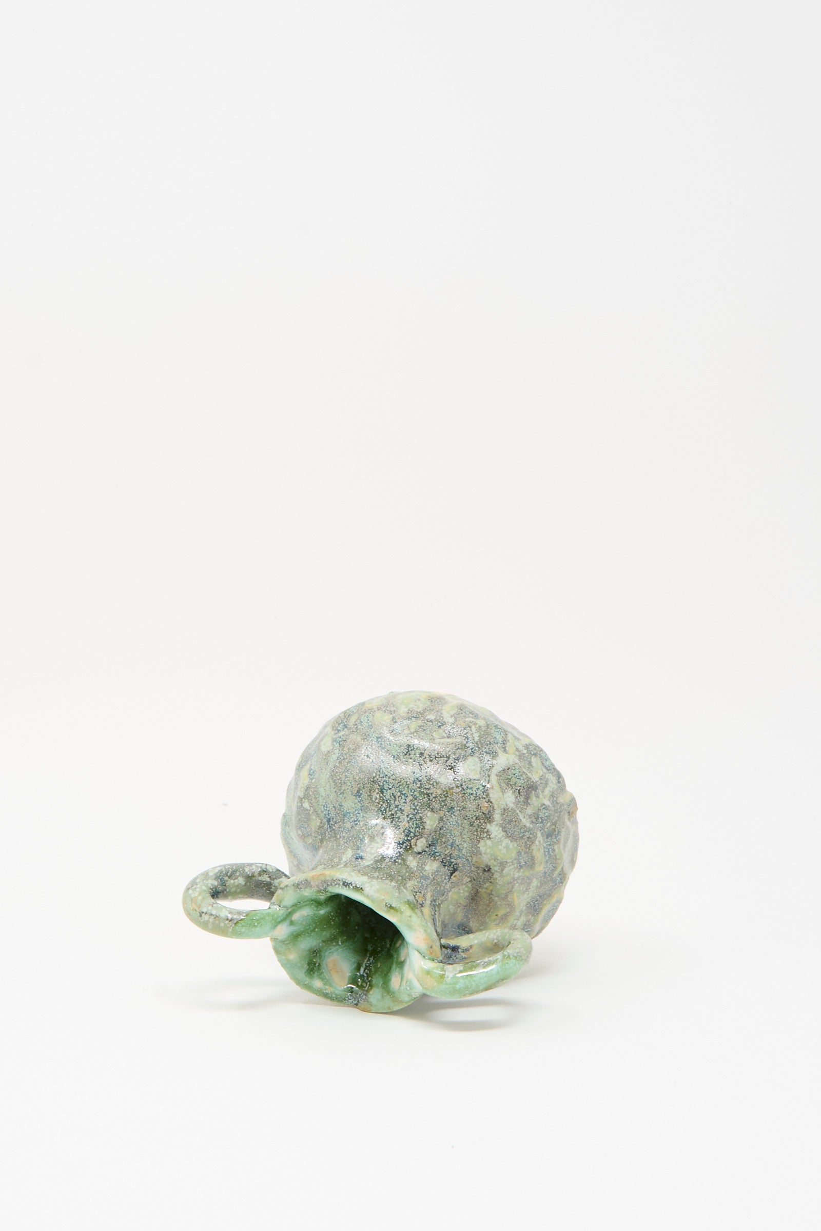 The MONDAYS Pinched Vase with Handles in Green, a small ceramic piece with a textured surface and two side handles, is lying on its side against a plain white background.
