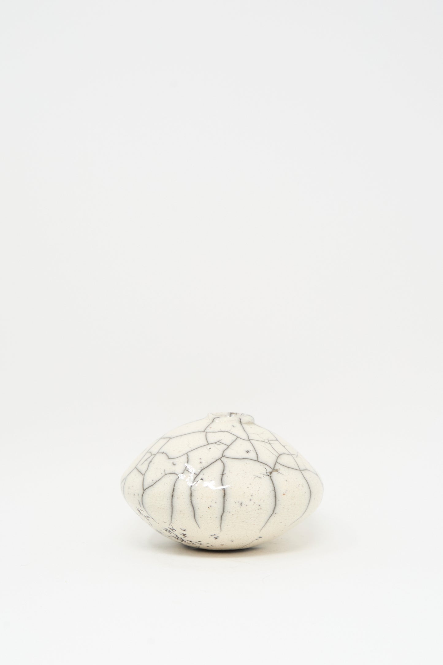 The MONDAYS Raku Moon Vase in White is a small, round ceramic piece with dark, irregular crackle lines, set against a plain white background.