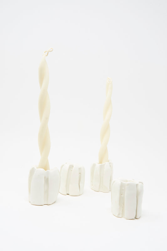 Two twisted white candles in sculptural handmade porcelain MONDAYS candleholders, including an empty MONDAYS Ribbed Candleholder in White, all set against a plain white background.