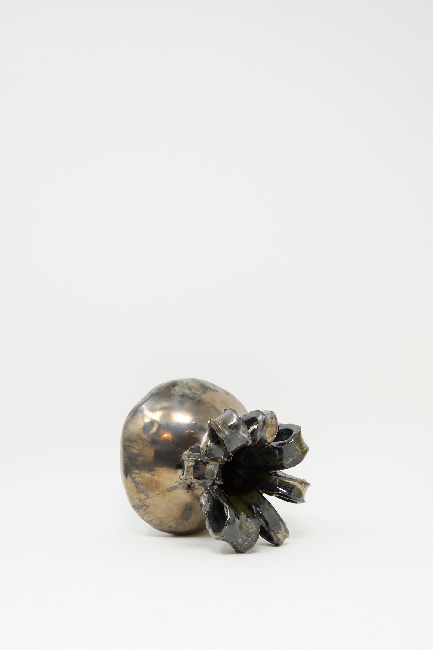 The Ribbon Vase in Mirror by MONDAYS, featuring a metallic silver finish and sculpted to resemble a poppy seed pod, rests on its side against a plain white background.