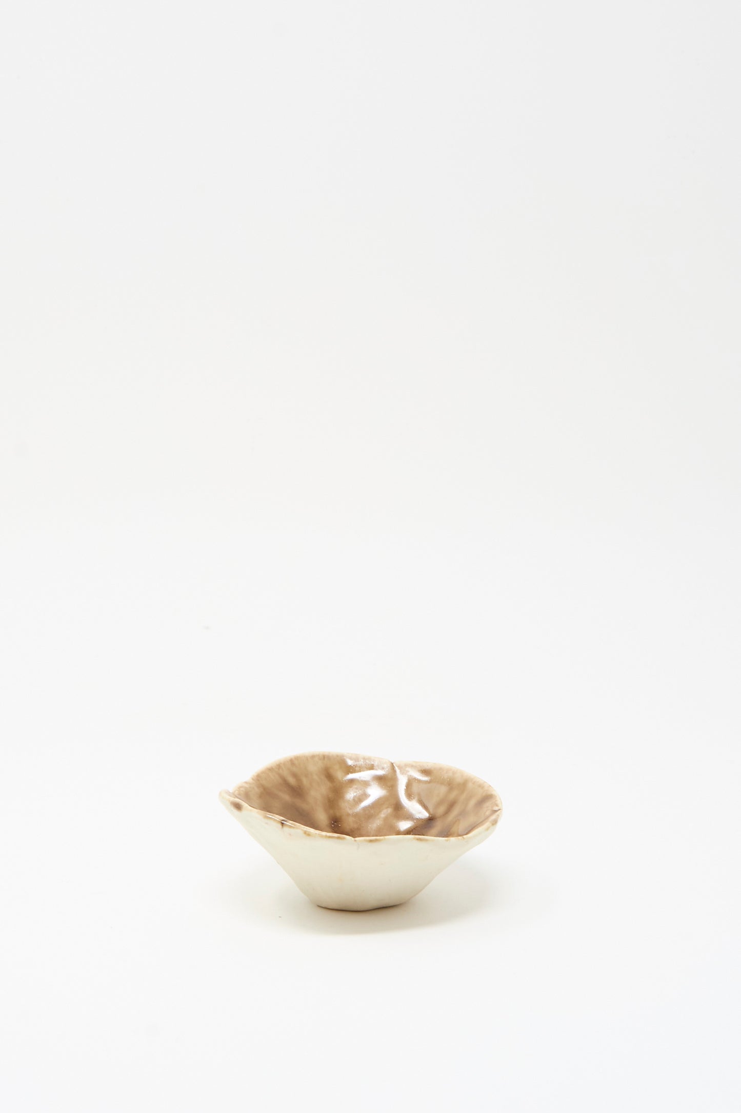 The Small Pinched Bowl in Brown by MONDAYS features a shallow, handmade design with an irregular rim and glossy light brown glaze, shown against a plain white background.