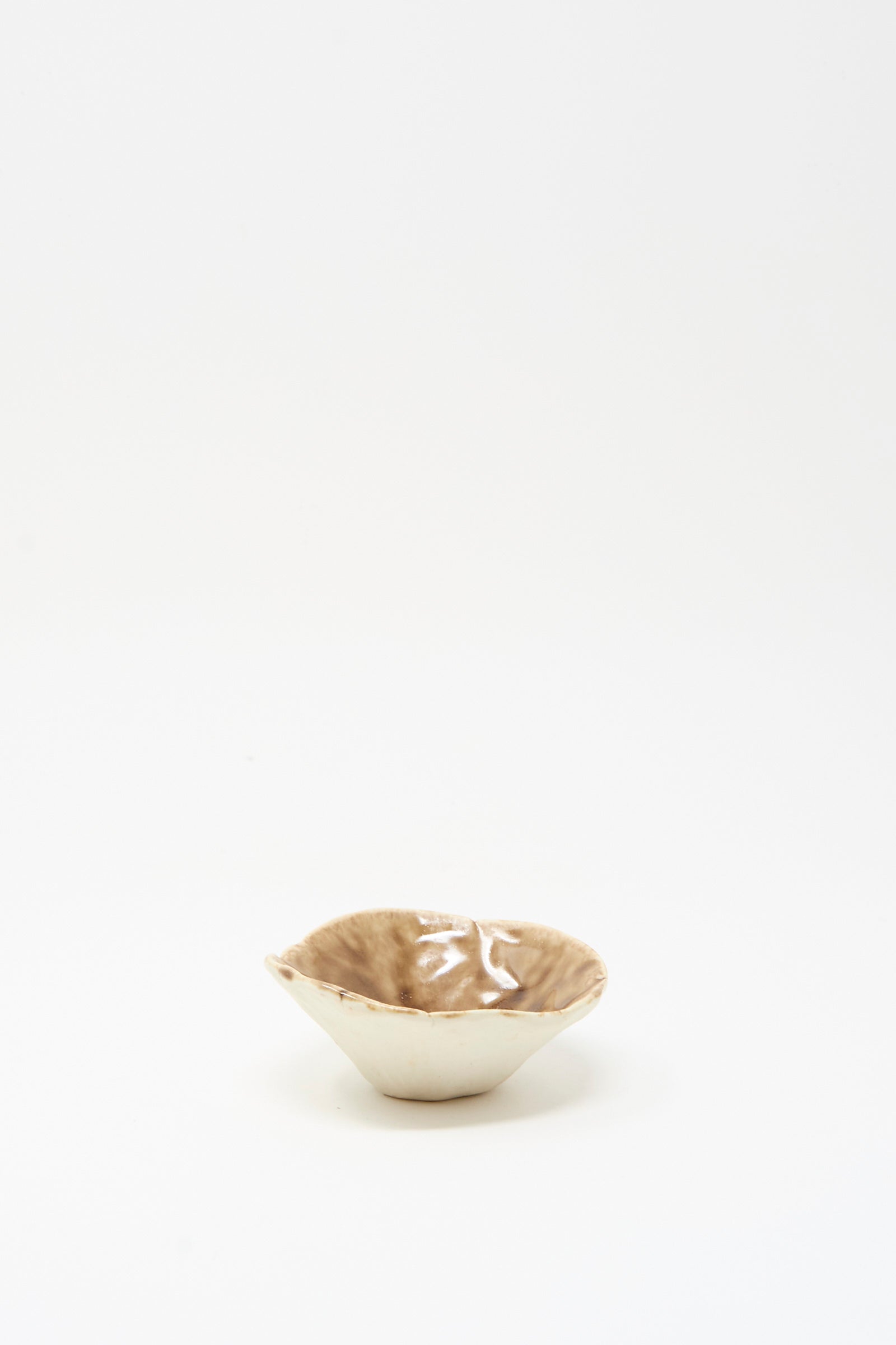 The Small Pinched Bowl in Brown by MONDAYS features a shallow, handmade design with an irregular rim and glossy light brown glaze, shown against a plain white background.