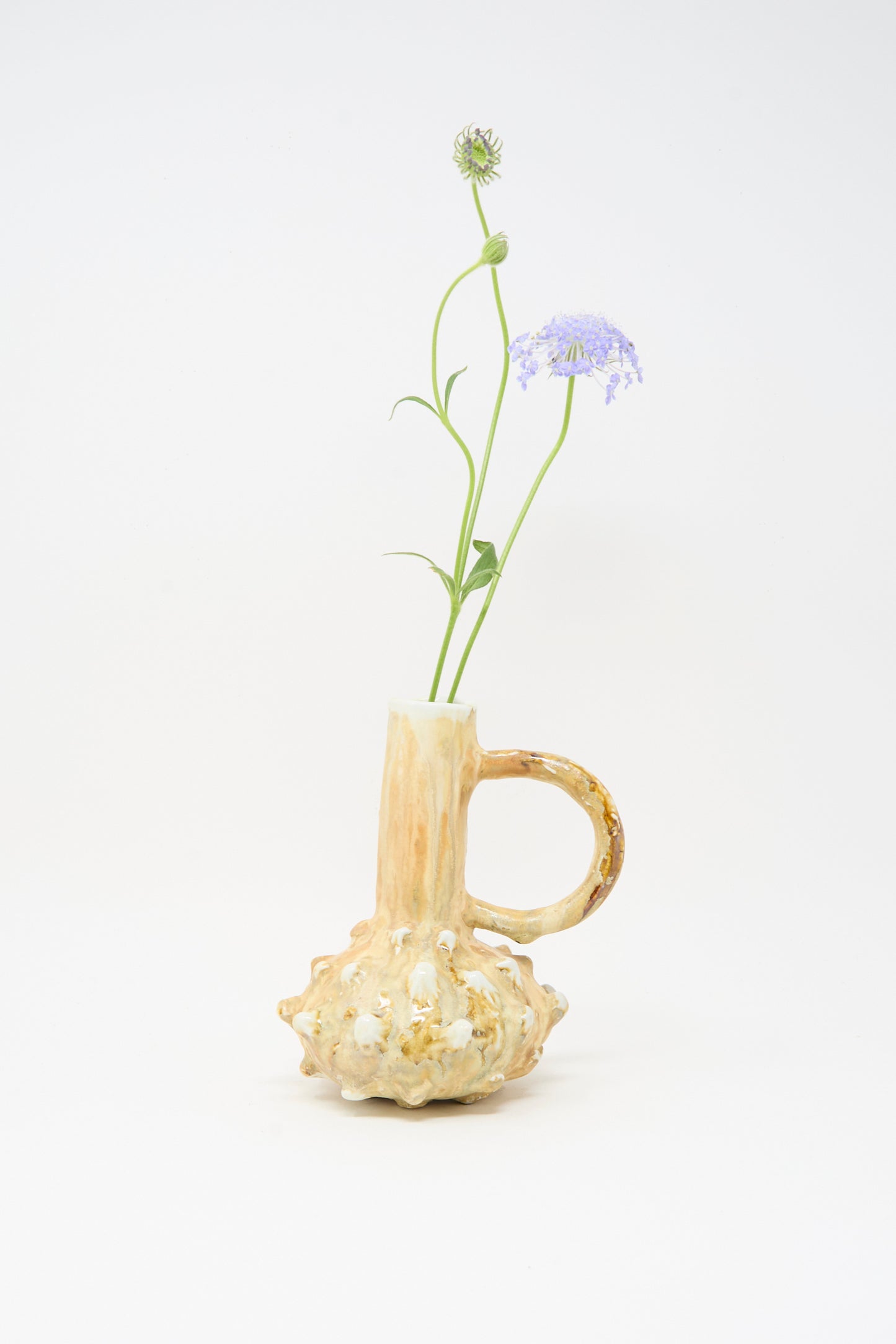 The MONDAYS Soda-Fired Bottle Vase No. 7 features a textured, bulbous beige ceramic base and holds two long-stemmed purple flowers against a plain white background.