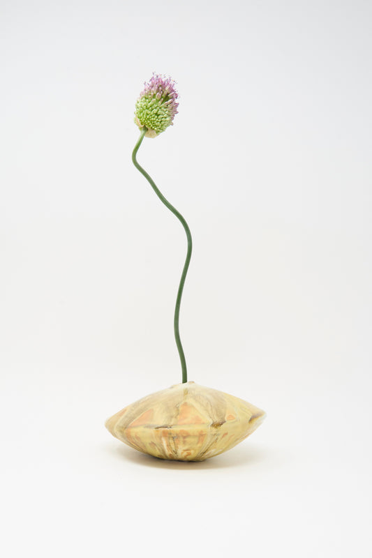 A single purple allium with a curved green stem in the MONDAYS Soda-Fired Ikebana Vase No. 1, a round handmade vase crafted from beige ceramic, displayed against a plain white background.