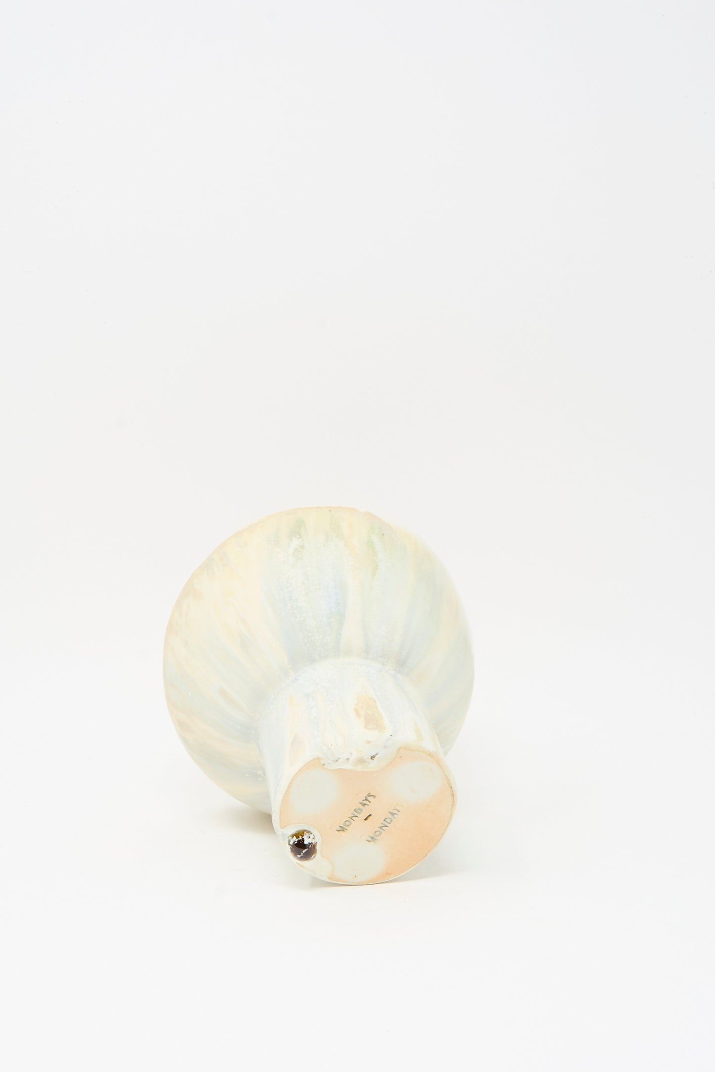 The MONDAYS Soda-Fired Pedestal Bowl, handmade in porcelain with a rounded top and flat base, lies on its side next to a small metallic ball on a white background.