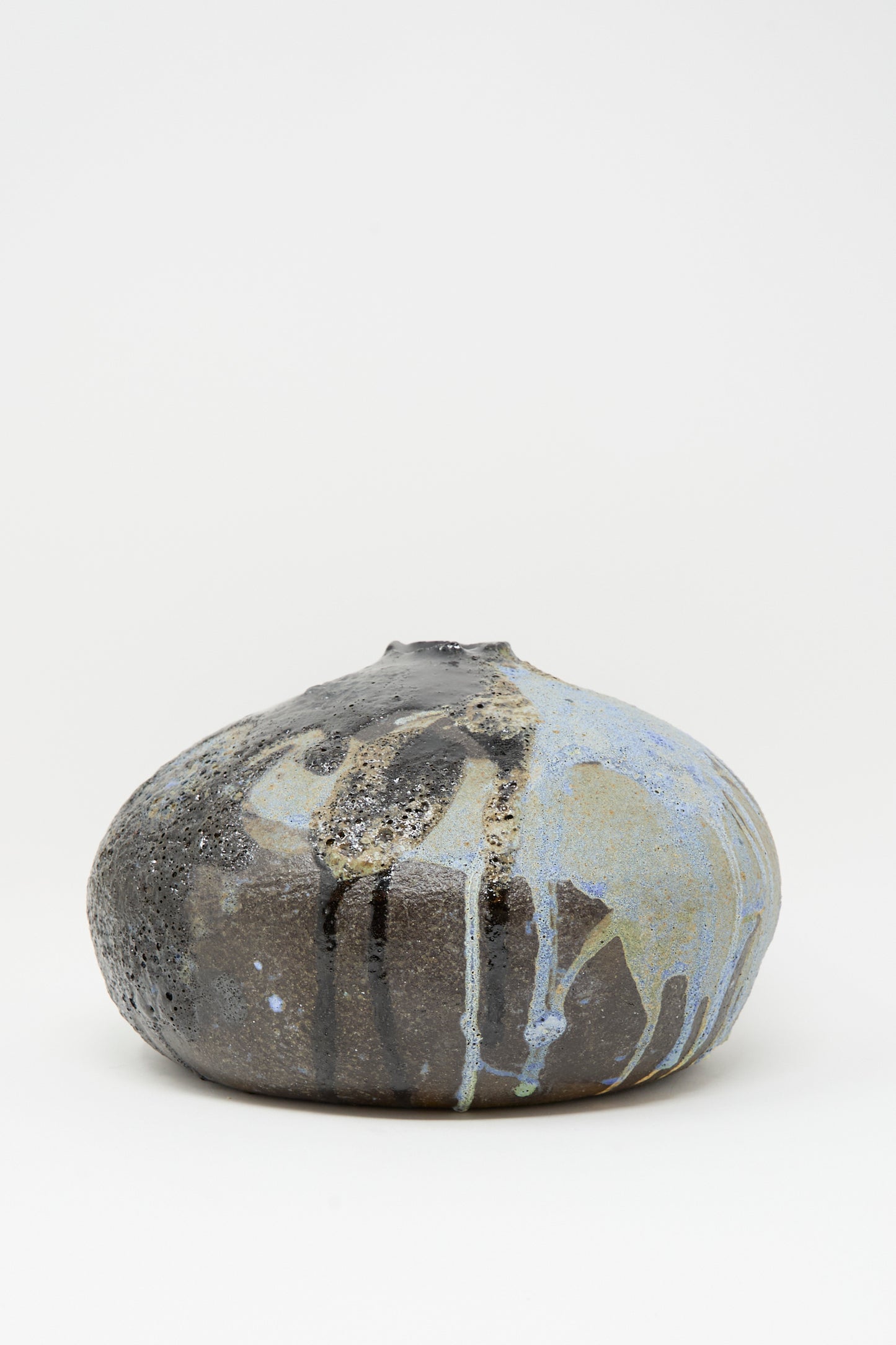 The MONDAYS Textured Moon Vase in Black is a handmade round vase with a small opening, featuring a dark textured surface and multicolored blue, white, and gray glaze drips that stand out against a plain white background.