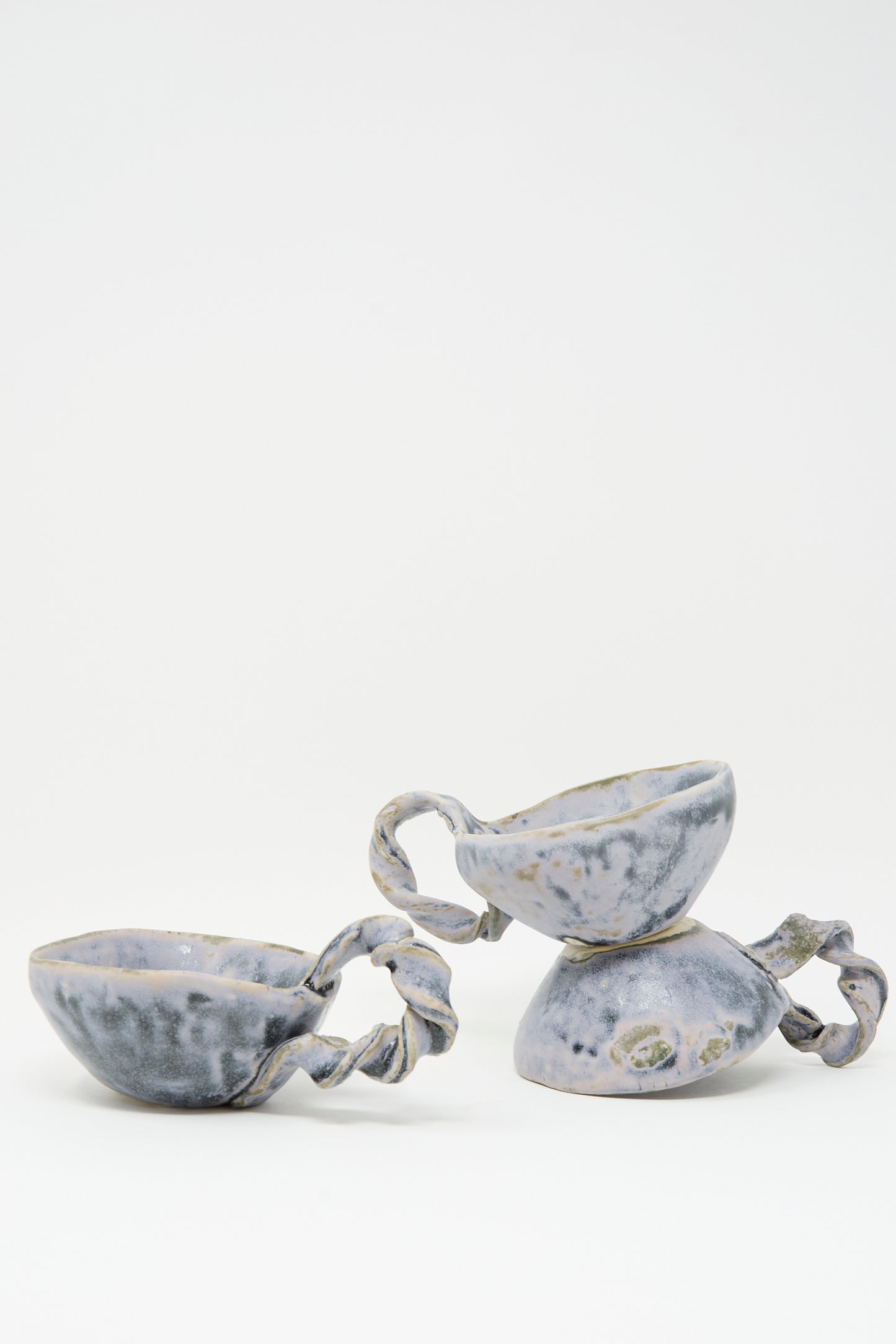The MONDAYS Twist Teacup in Blue features handmade porcelain with twisted handles and a blue-gray speckled glaze on white. Thoughtfully crafted in Brooklyn, it comes as a set of two—the cups shown upright and leaning on their side.