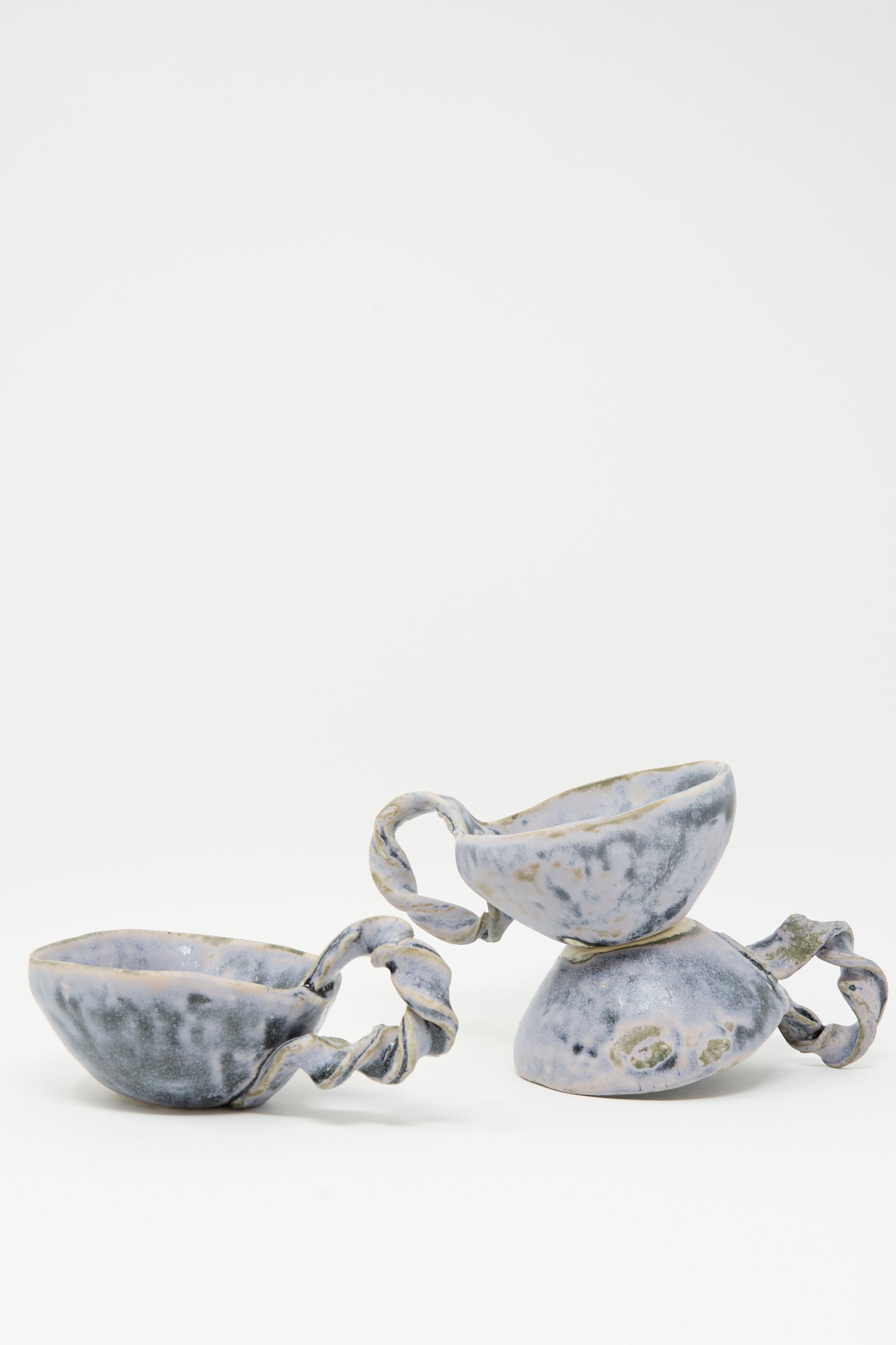 The MONDAYS Twist Teacup in Blue features handmade porcelain with twisted handles and a blue-gray speckled glaze on white. Thoughtfully crafted in Brooklyn, it comes as a set of two—the cups shown upright and leaning on their side.