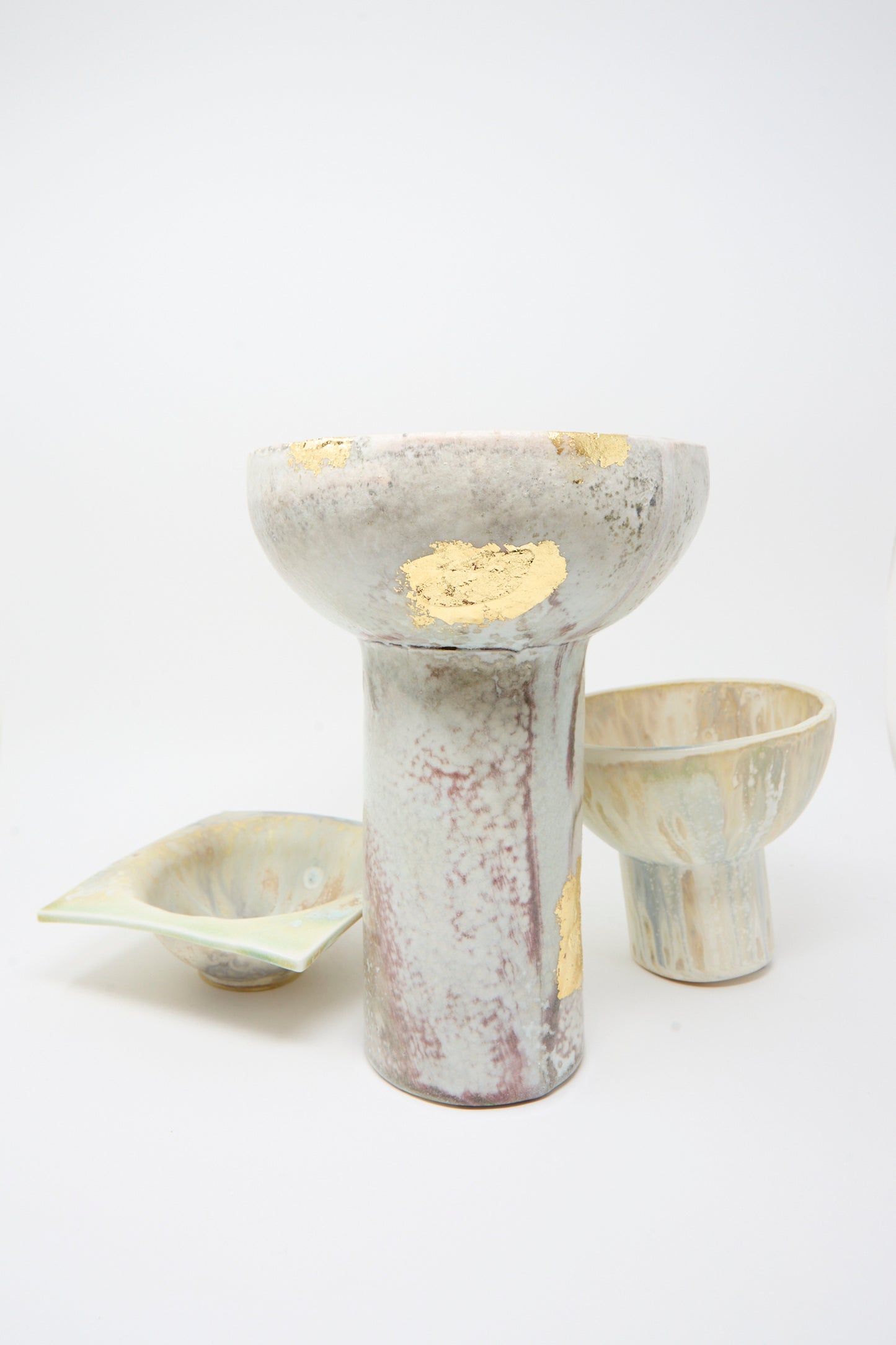 Three ceramic vessels with white and marbled glaze and gold leaf accents sit against a plain backdrop. Featured is the MONDAYS Wood-Fired Porcelain Pedestal Bowl with Gold Leaf, showcasing gold detailing.