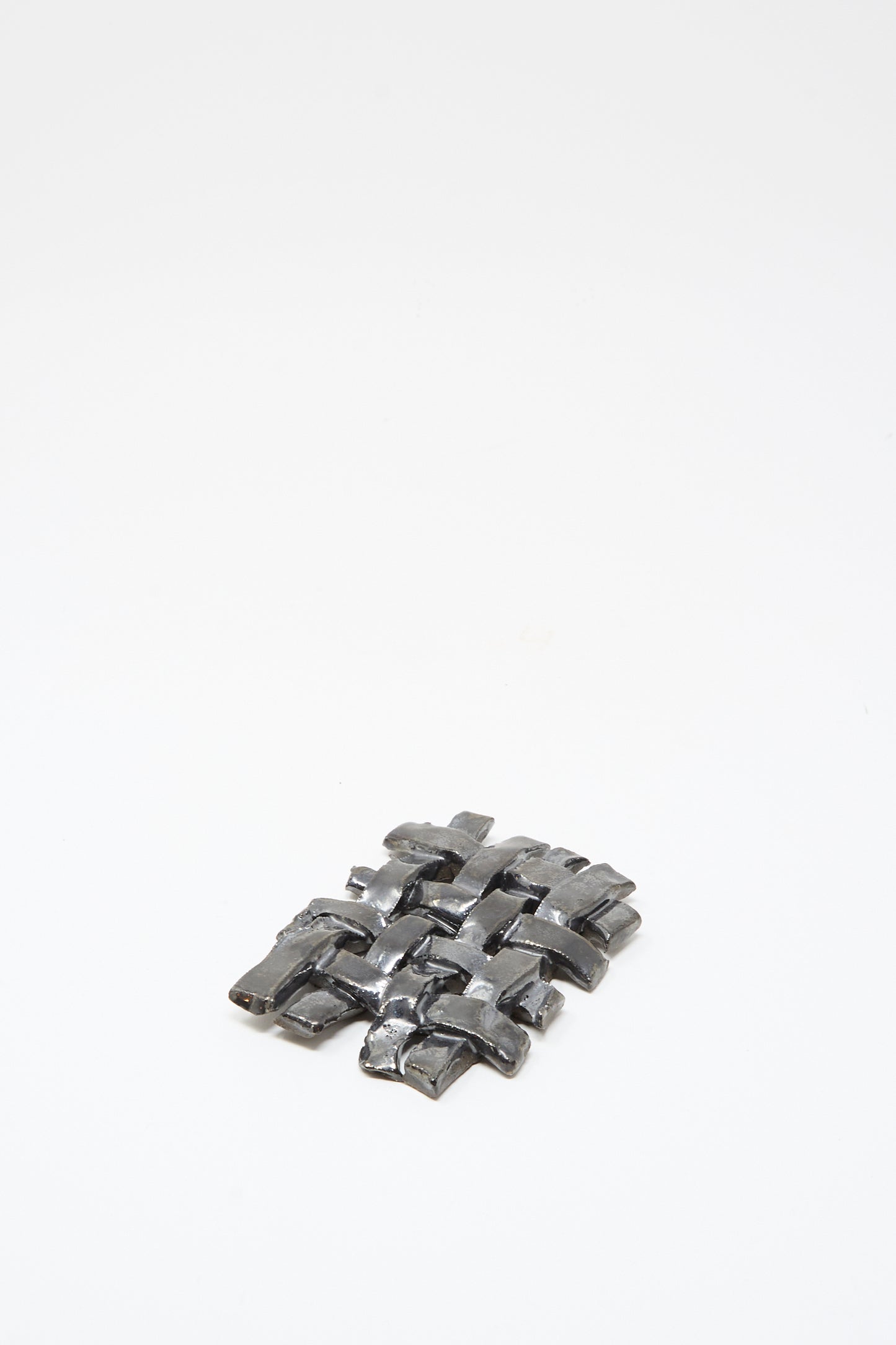 A small woven grid of shiny metal strips on a white background, resembling the MONDAYS Woven Dish in Charcoal.
