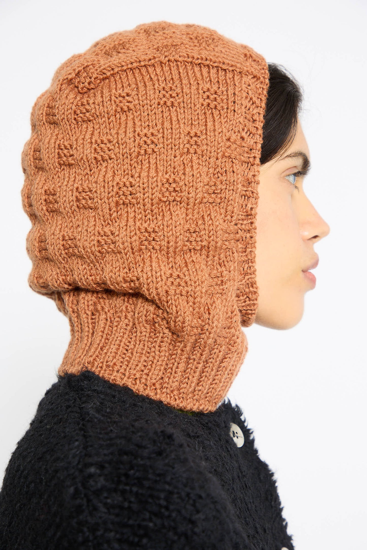 Side profile of a person wearing the Balaclava in Rust, hand-knit by Maria La Rosa, paired with a black garment. The background is plain white.