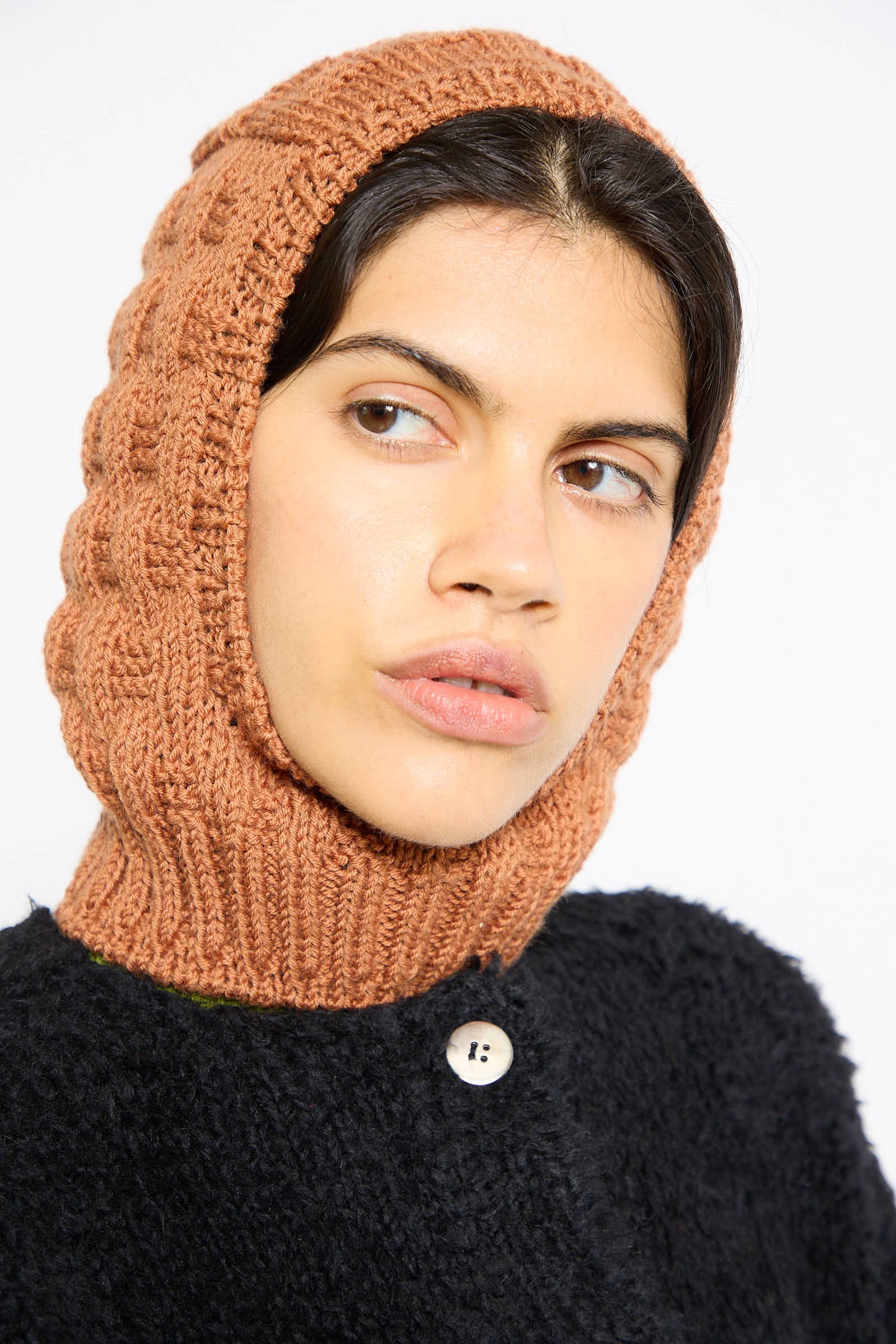 A person with dark hair is wearing the Maria La Rosa Balaclava in Rust and a black textured top with a button.