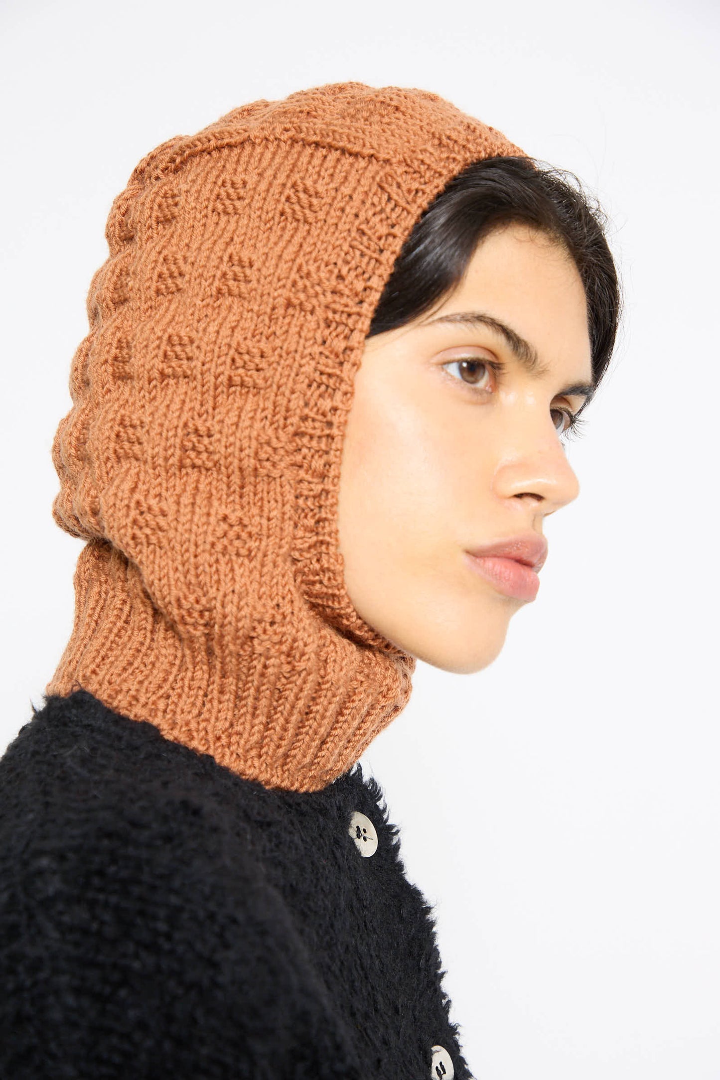 A person wearing a Maria La Rosa Balaclava in Rust and a black textured top with large buttons is pictured in profile against a plain background.