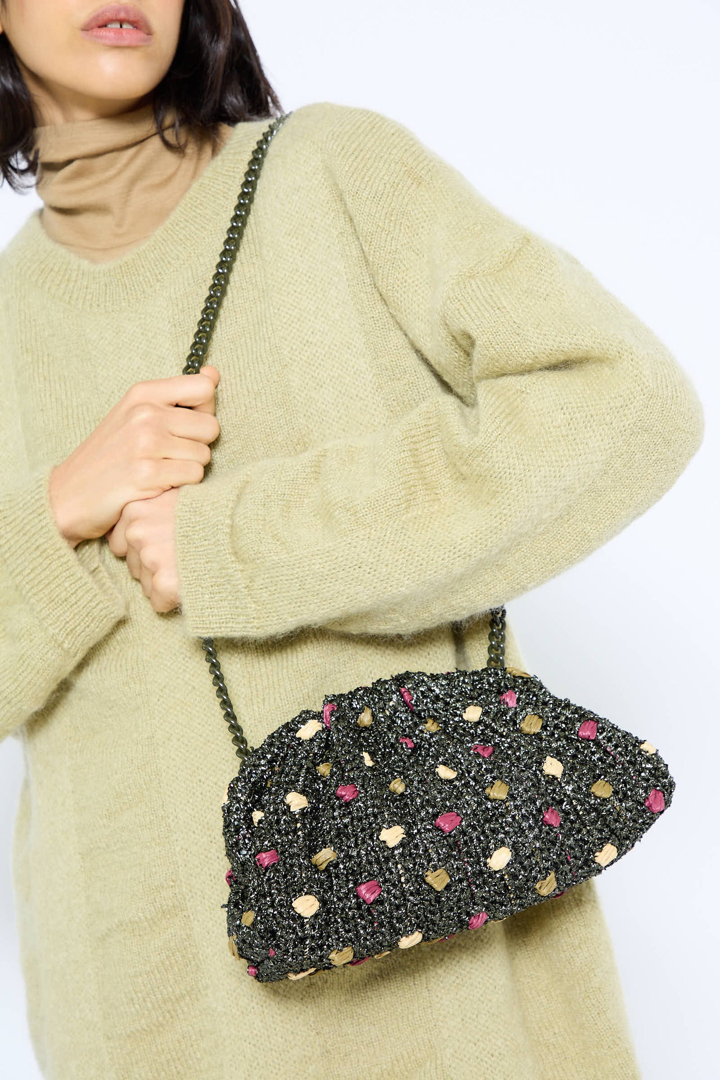 A person in a beige sweater holds the Candy Peanuts Game Bag in Military by Maria La Rosa, a black clutch adorned with delicate pink and yellow accents, featuring an elegant chain shoulder strap.