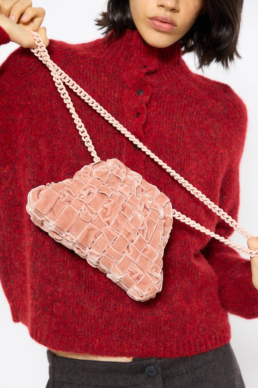 A person wearing a red knit sweater holds a Velvet Mini Game Bag in Cipria by Maria La Rosa, featuring a pink handwoven velvet design and a chain shoulder strap.