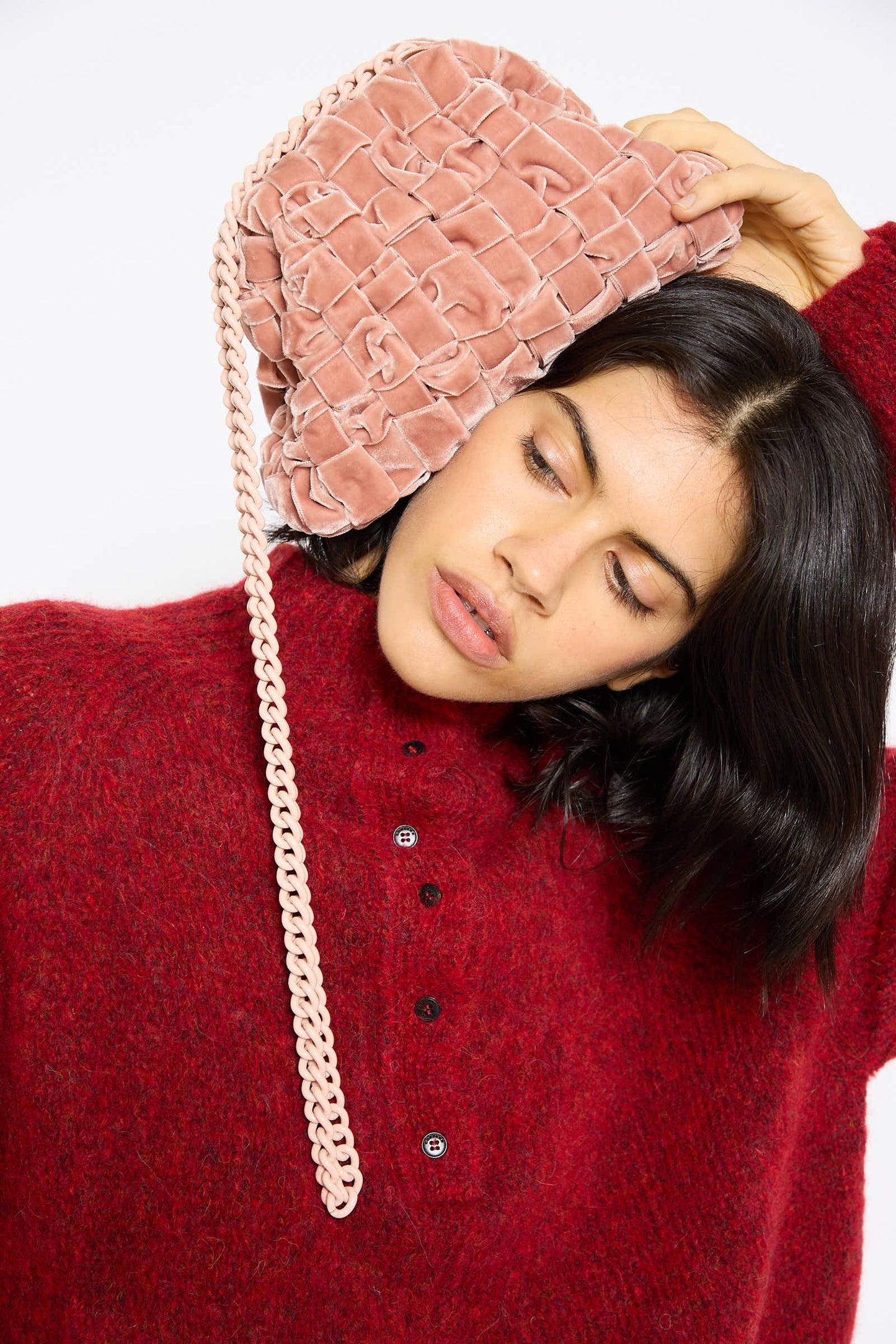 A person with black hair wearing a red sweater holds the Maria La Rosa Velvet Mini Game Bag in Cipria, a pink handwoven velvet handbag with a chain shoulder strap, against their head.