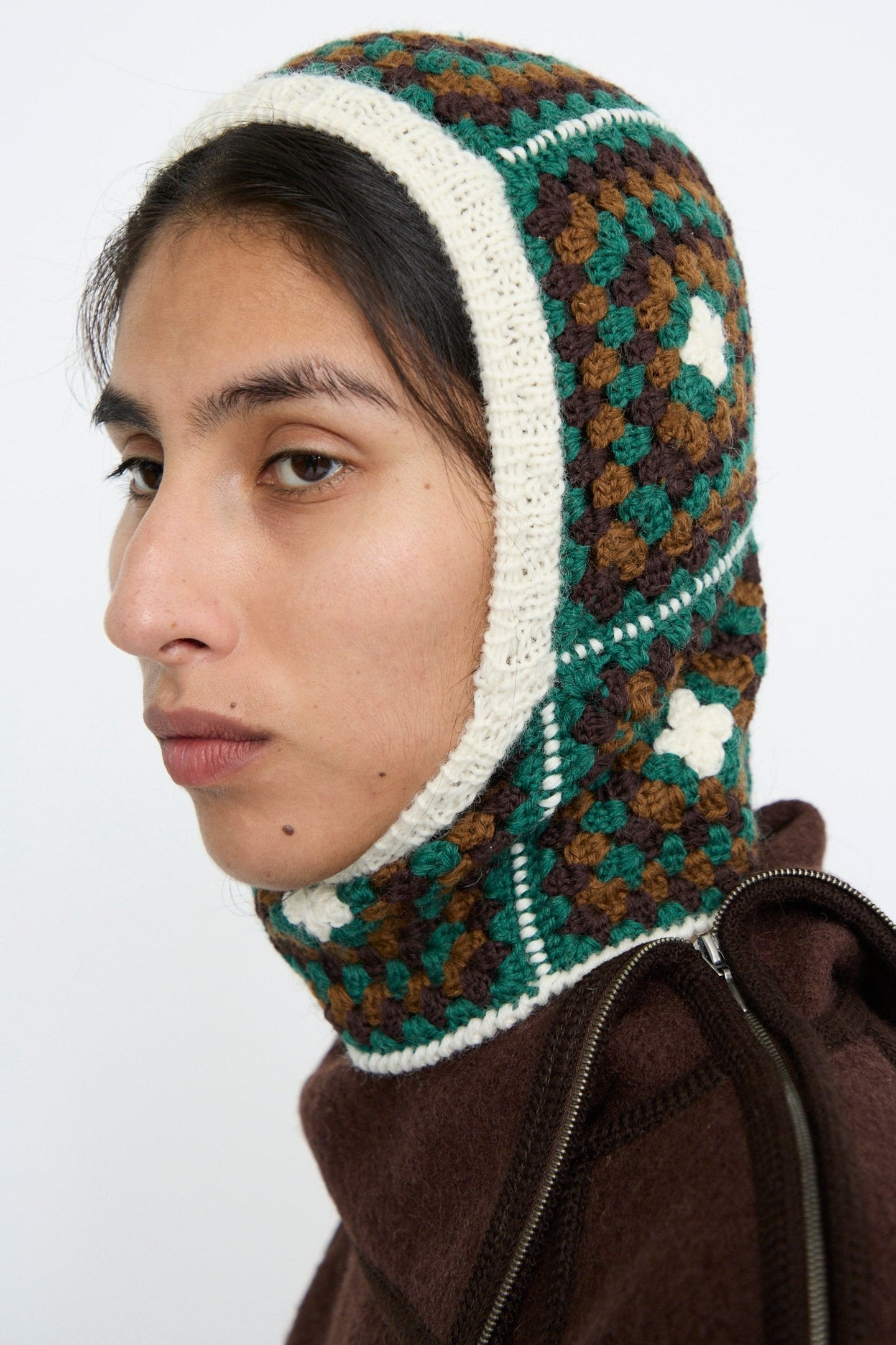 A person is seen wearing the Balaclava in Emerald by Maria La Rosa, showcasing a hand-knit look with a crocheted wool pattern of green, brown, and white squares in a multi-colored design from the side view.