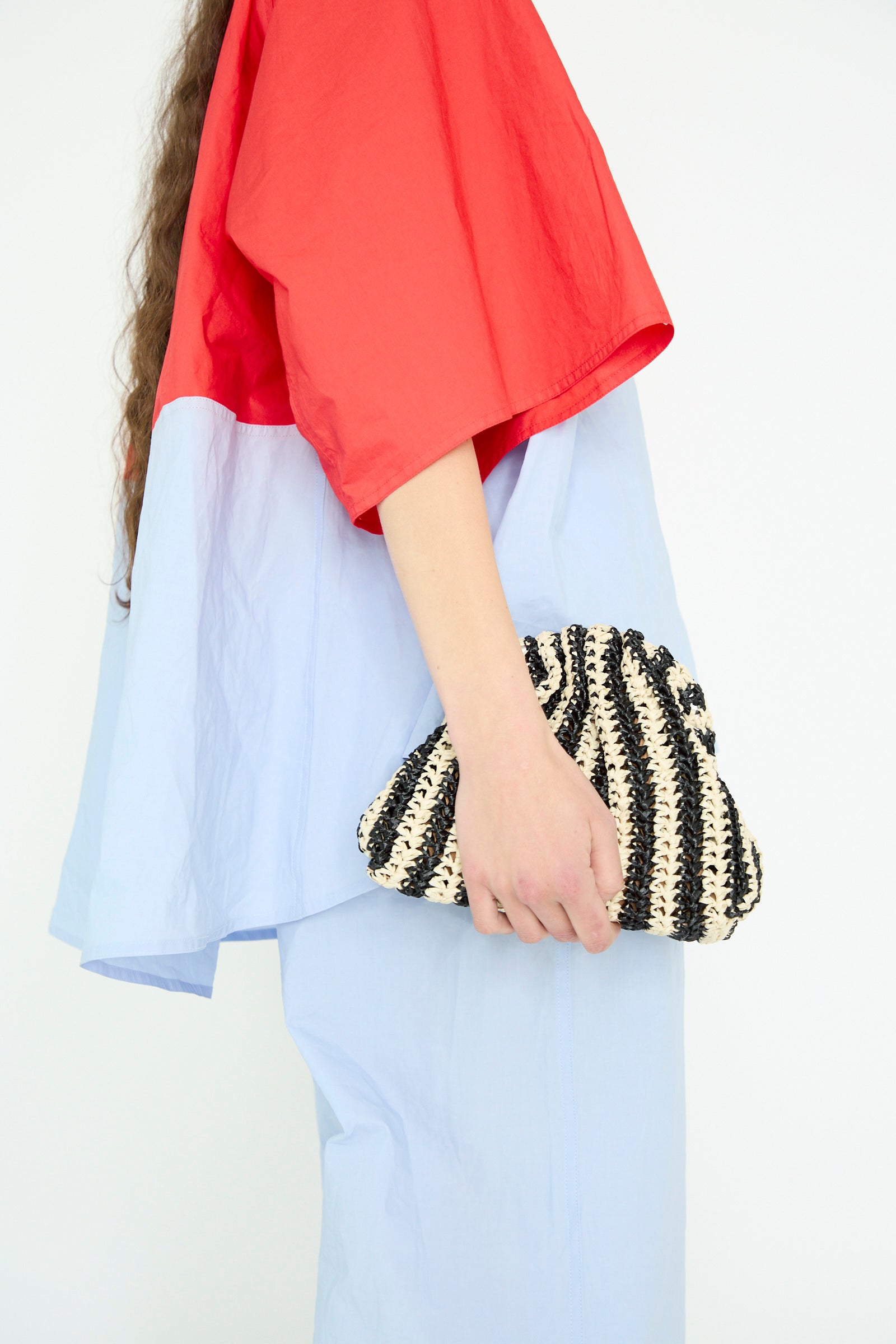 A person in a red and light blue outfit holds the Crochet Mini Game Cromo Bag by Maria La Rosa, a black-and-white striped mini clutch featuring a chain shoulder strap, against a plain background.