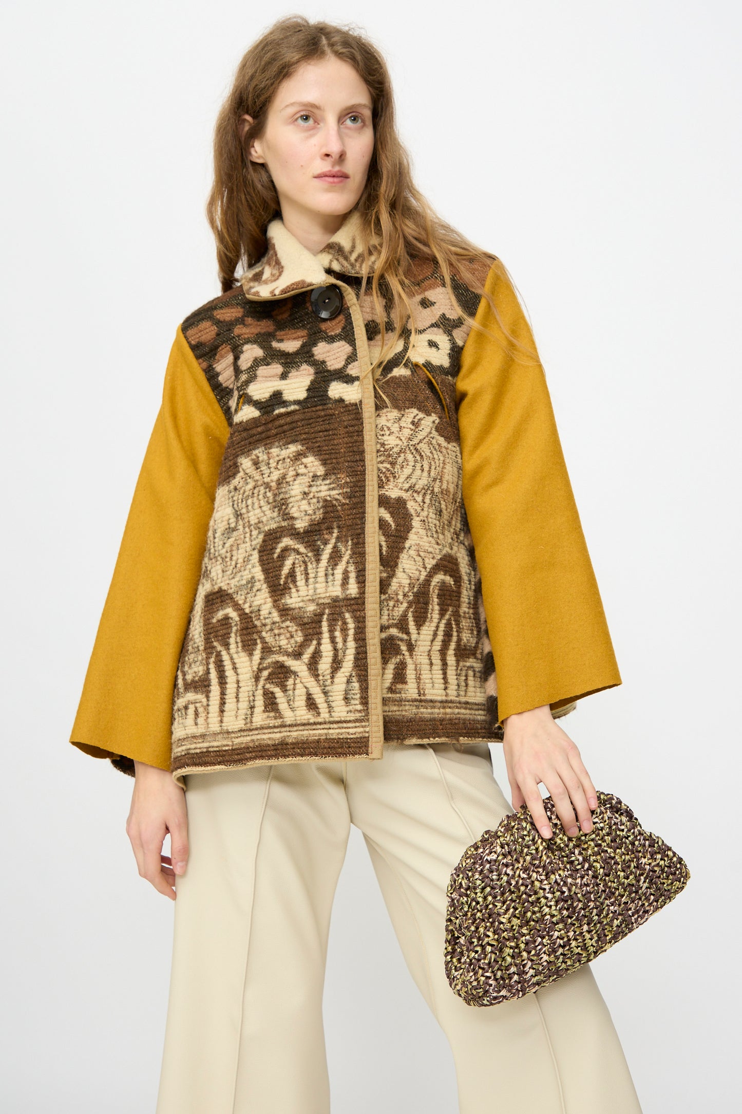 A person wearing a mustard yellow and brown patterned jacket with animal motifs, beige pants, and holding the Maria La Rosa Earth Game Crochet Bag in Natural stands against a plain white background.