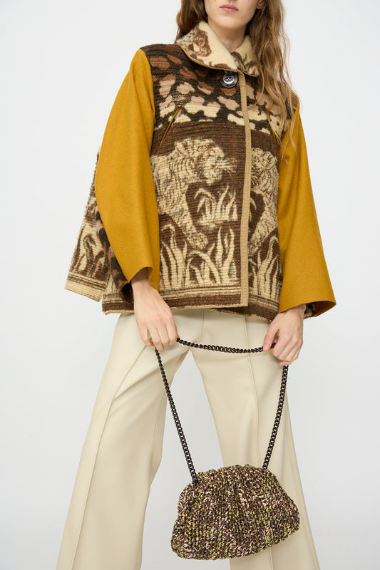 A woman in a patterned jacket with yellow sleeves and cream wide-leg pants holds the Maria La Rosa Earth Game Crochet Bag in Natural against a plain background.